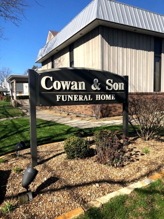Sign for Cowan & Son Funeral Home. Black sign with white text, in front of a tan building under a blue sky.