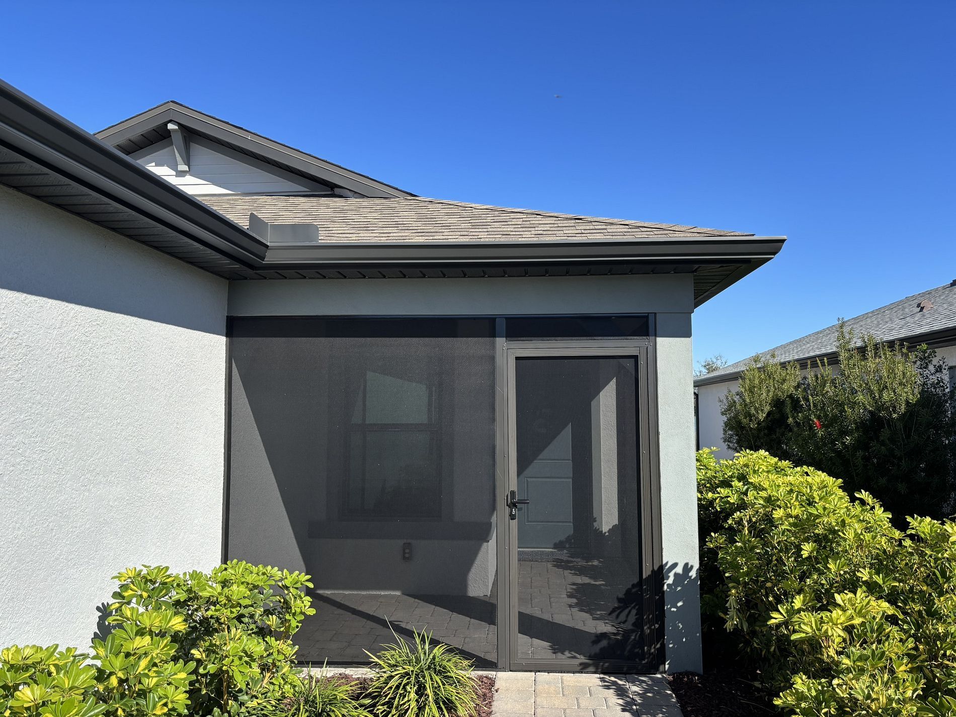 Screened-in porch with door, light blue exterior walls, gray roof, and green bushes against a clear blue sky.