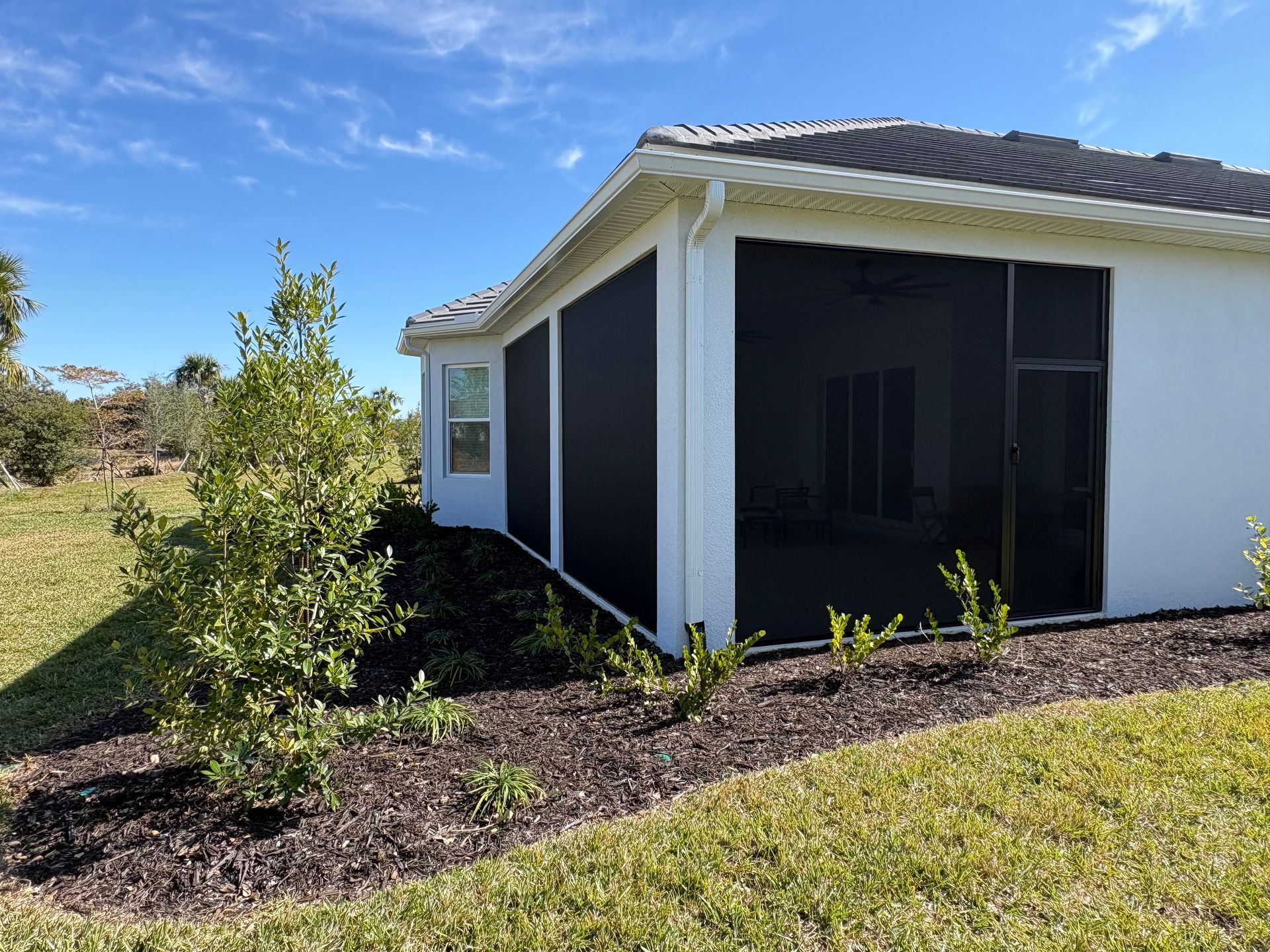 A white house with a screened-in patio, black screens, and landscaping under a blue sky.