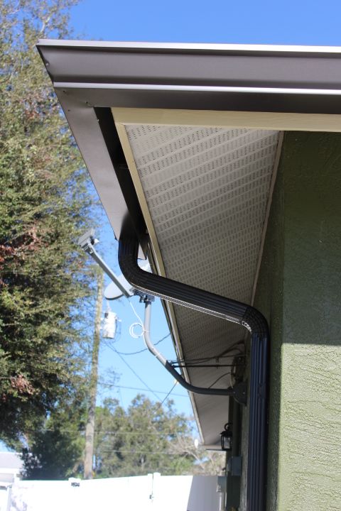 Black gutter and downspout on a light green house with white soffit, blue sky background.