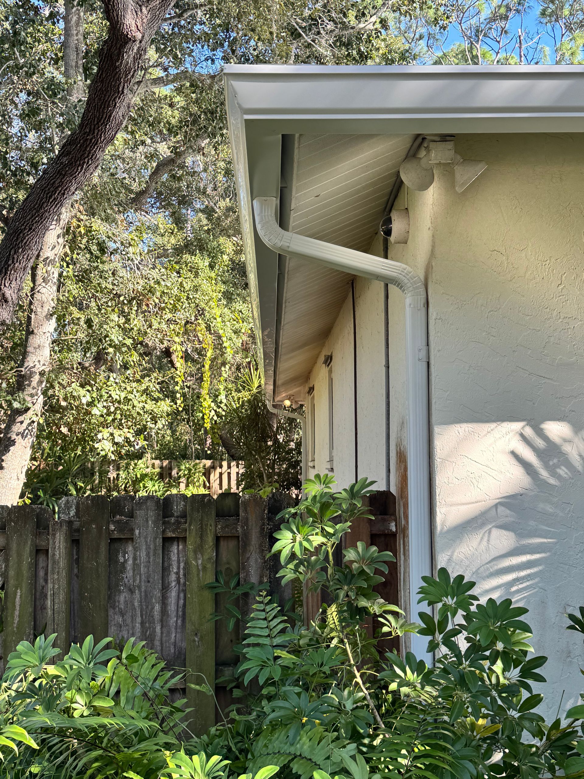 Corner of a house with white gutters and downspout; green foliage and a wooden fence are in the background.