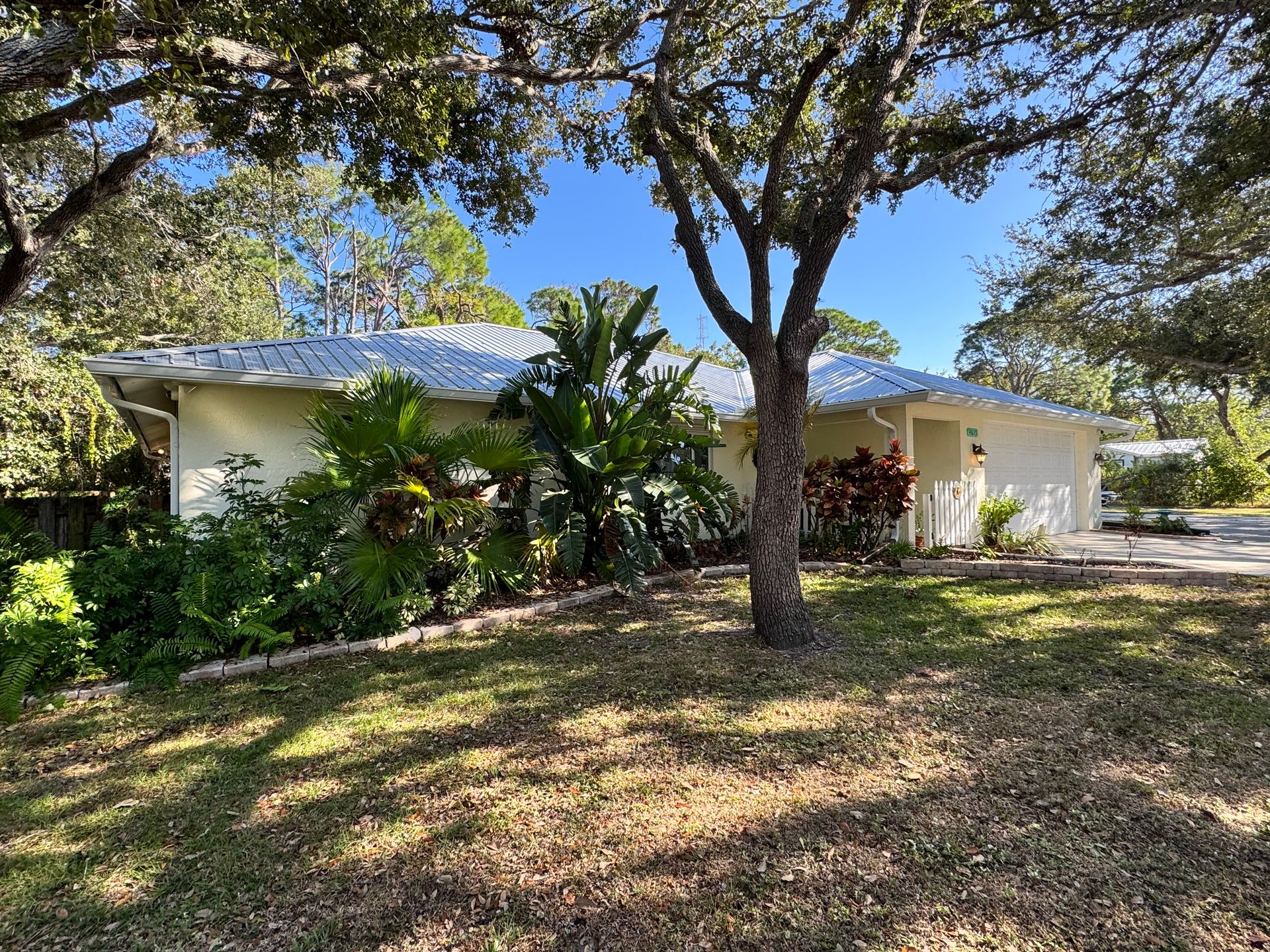Tan house with a light blue roof, surrounded by green foliage and trees under a clear sky.