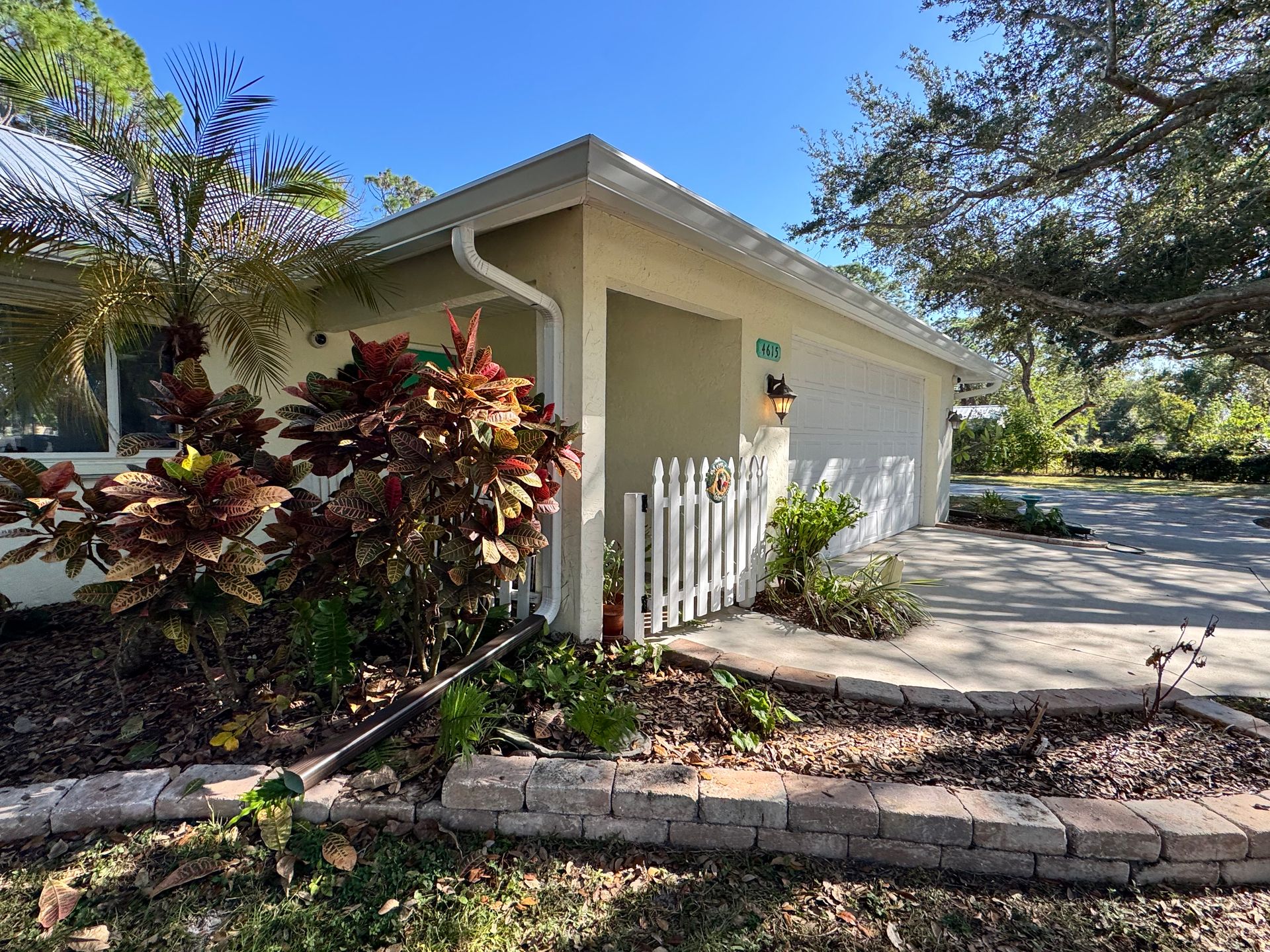 Beige house with a white picket fence, flower beds, and a driveway.