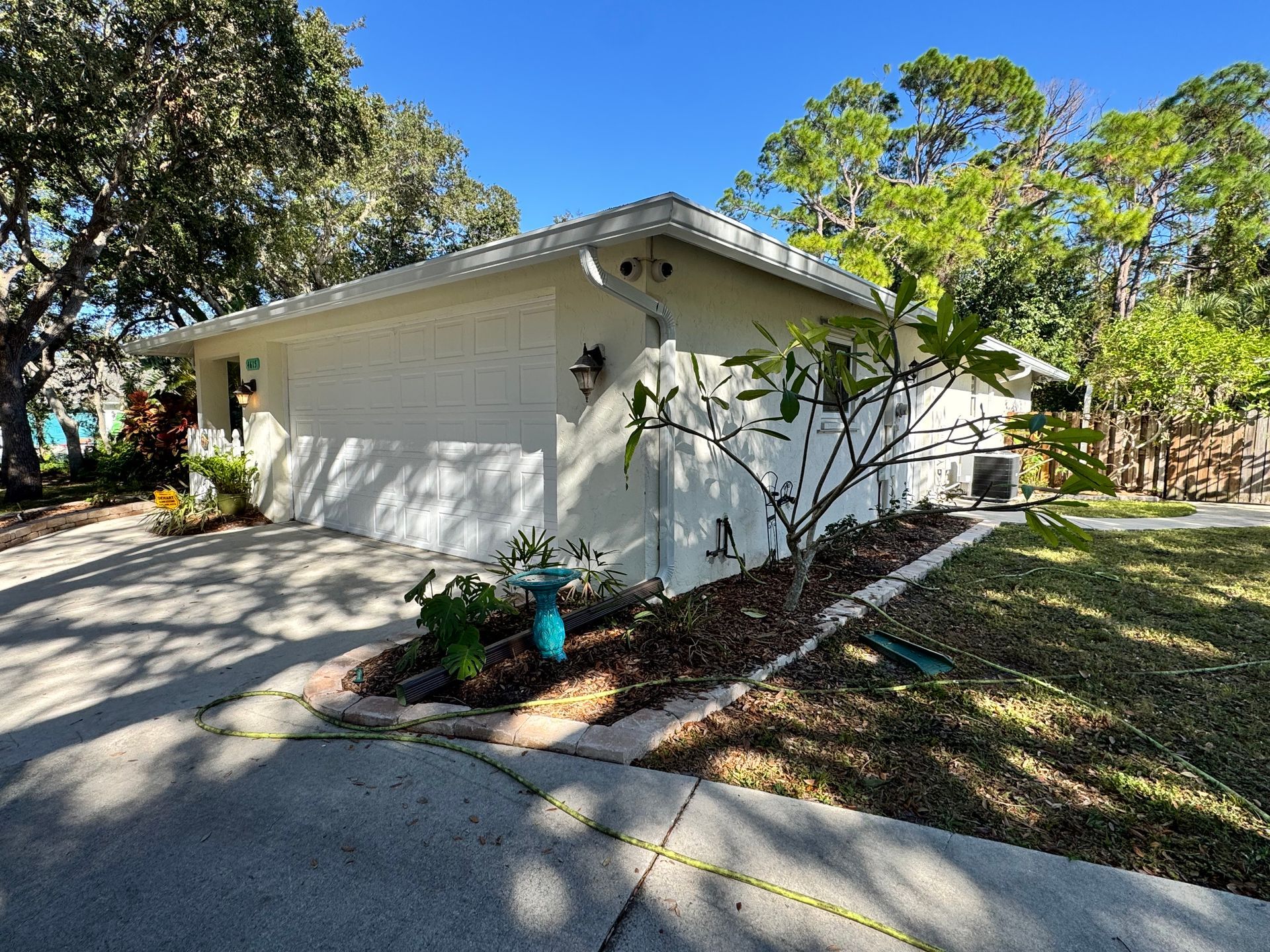 Light-colored house with attached garage, landscaped yard, and driveway under a clear blue sky.