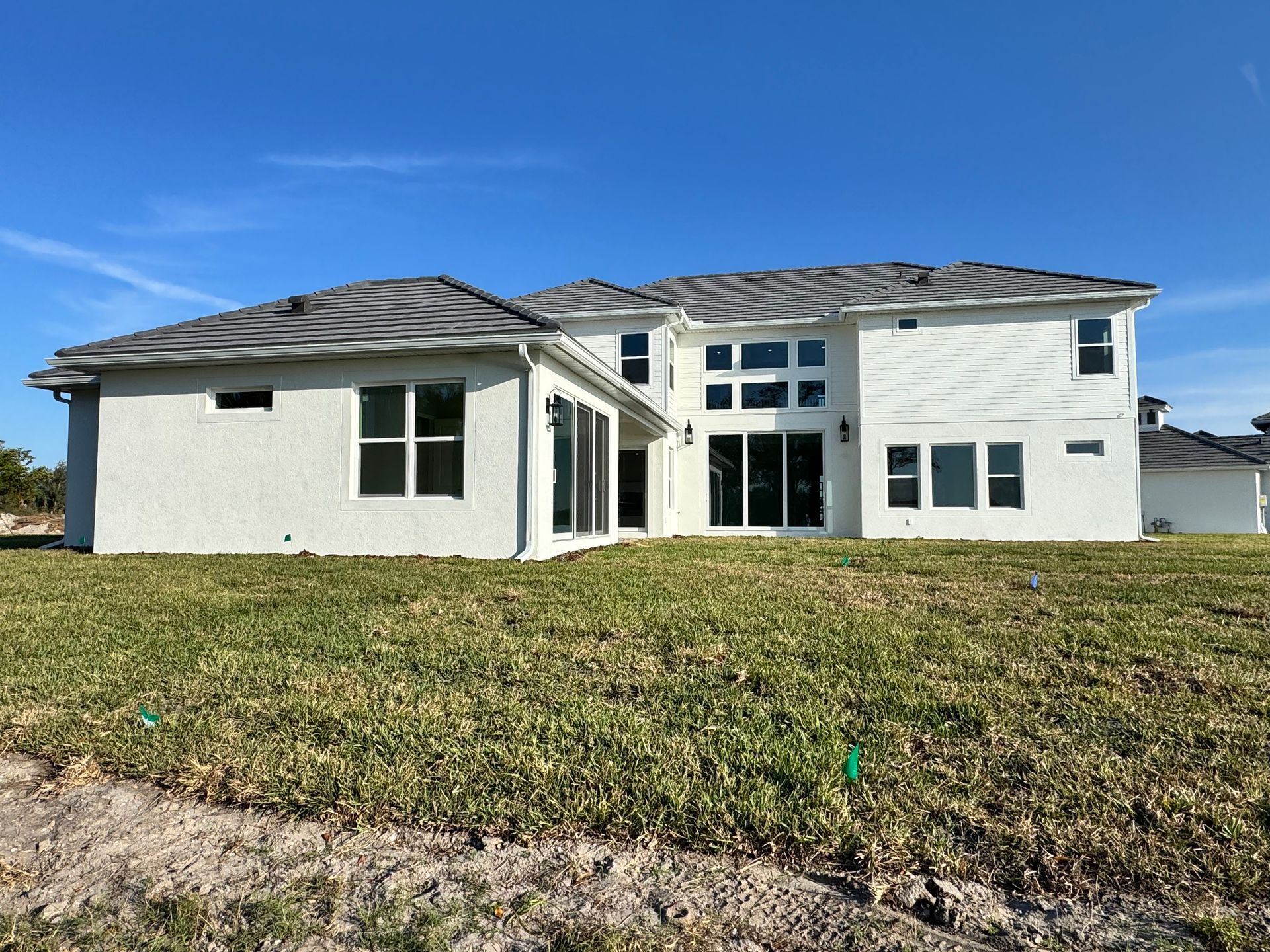 White two-story house with gray roof, large windows, and sliding glass doors. Situated on a grassy hill under a blue sky.