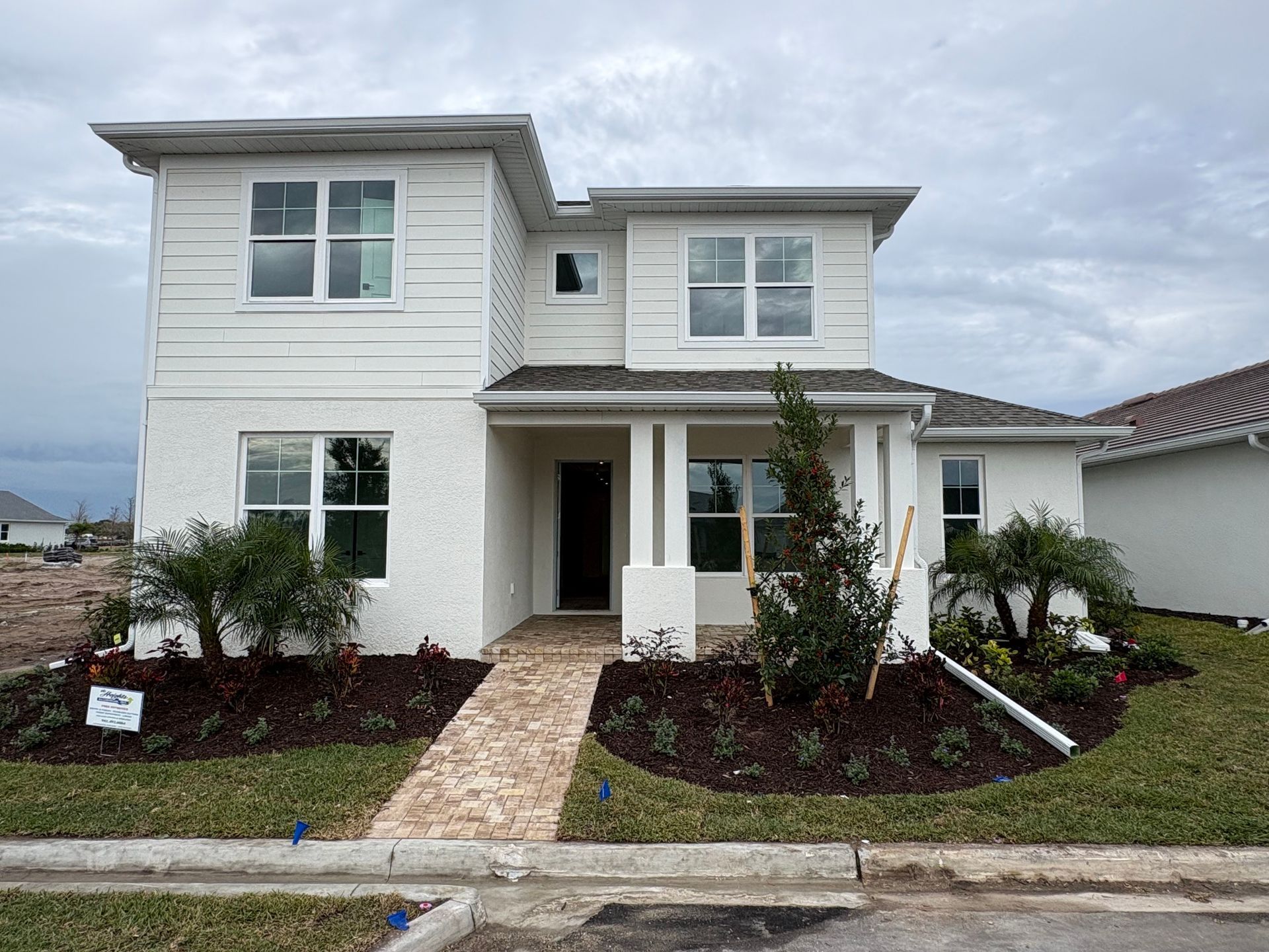 Two-story white house with brick walkway, small bushes, and landscaping under a cloudy sky.