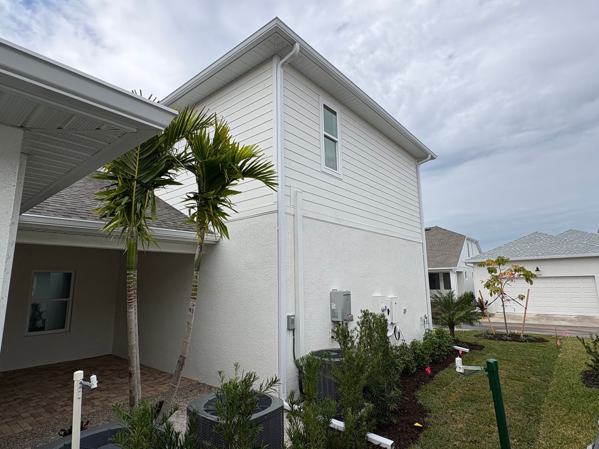 Two-story white house with a covered patio, palm trees, and green bushes. Overcast sky.