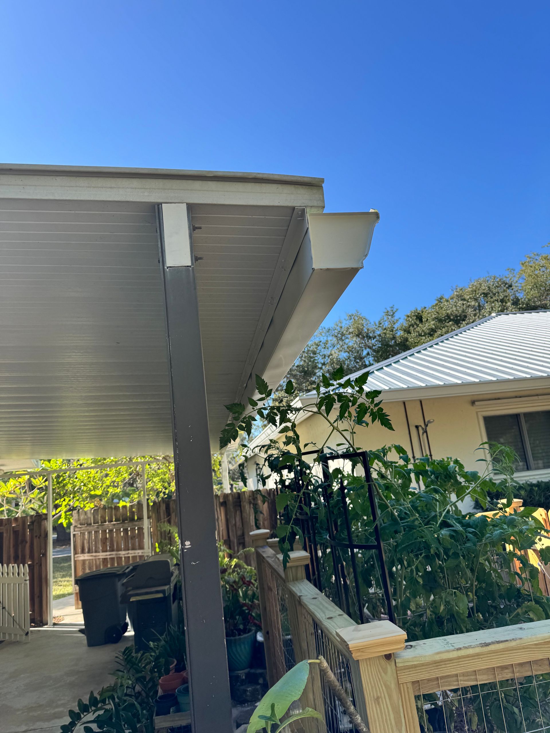 A covered patio with a white roof, gutter, and support posts next to a house with a blue sky.