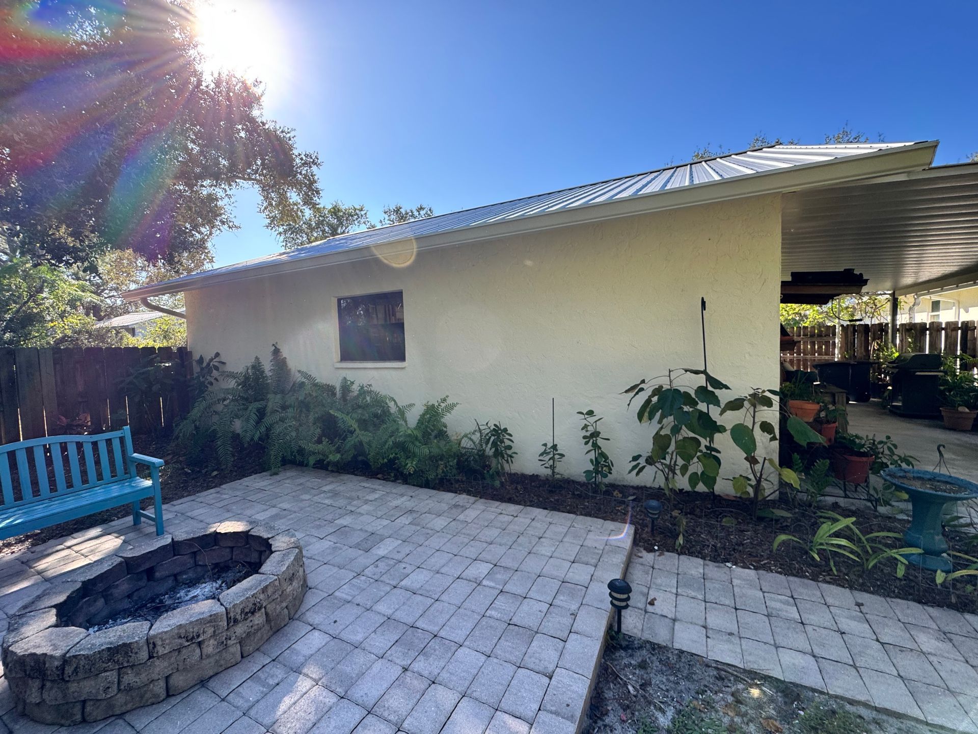 Backyard patio with fire pit, blue bench, plants, and a light-colored building under a sunny sky.