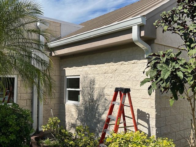 A house exterior with gutters, a ladder, and surrounding greenery under a blue sky.