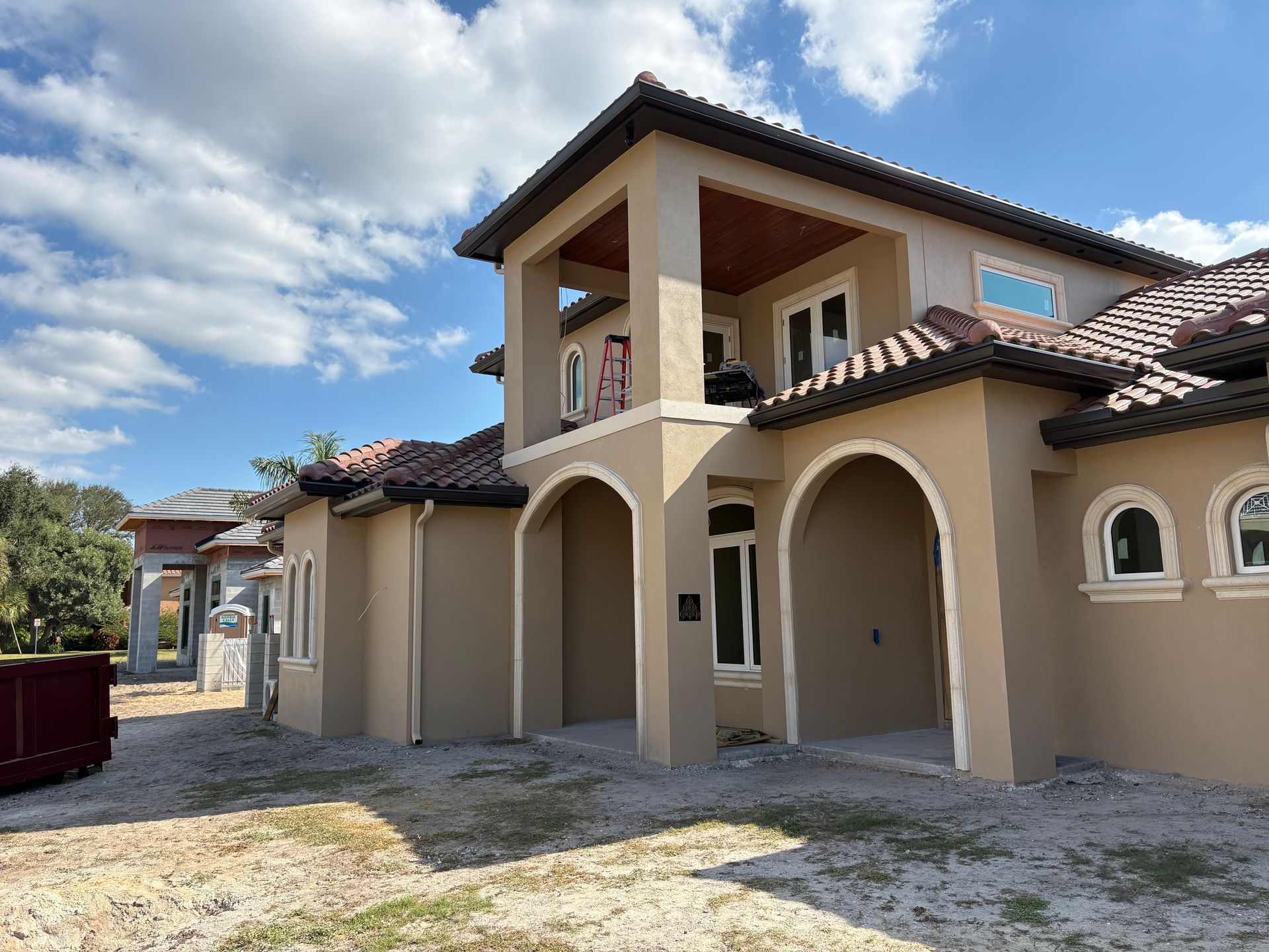 Tan stucco house with red-tile roof and second-story balcony under a cloudy blue sky.