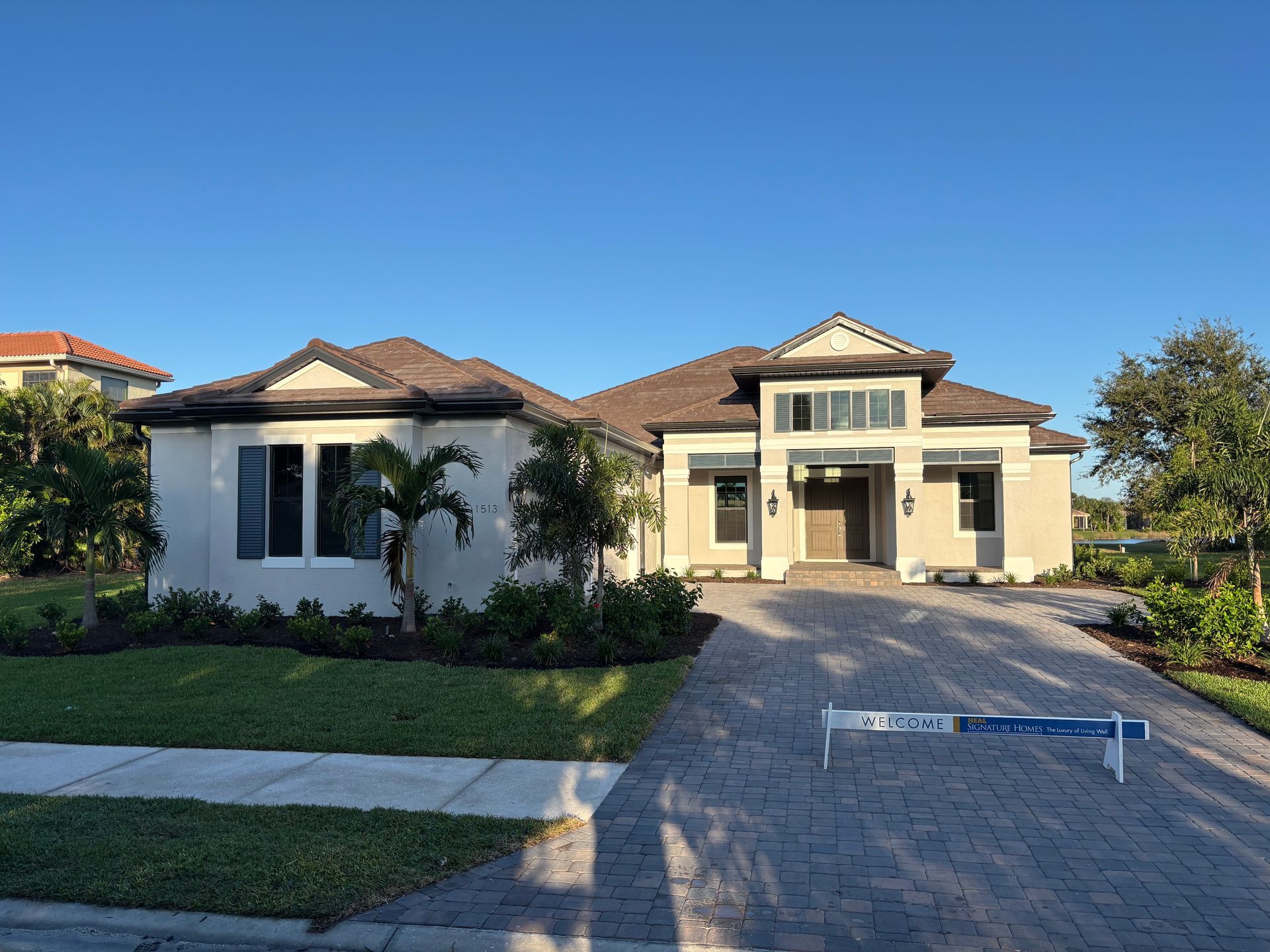 A light-colored, single-story house with a brick driveway and blue sky.