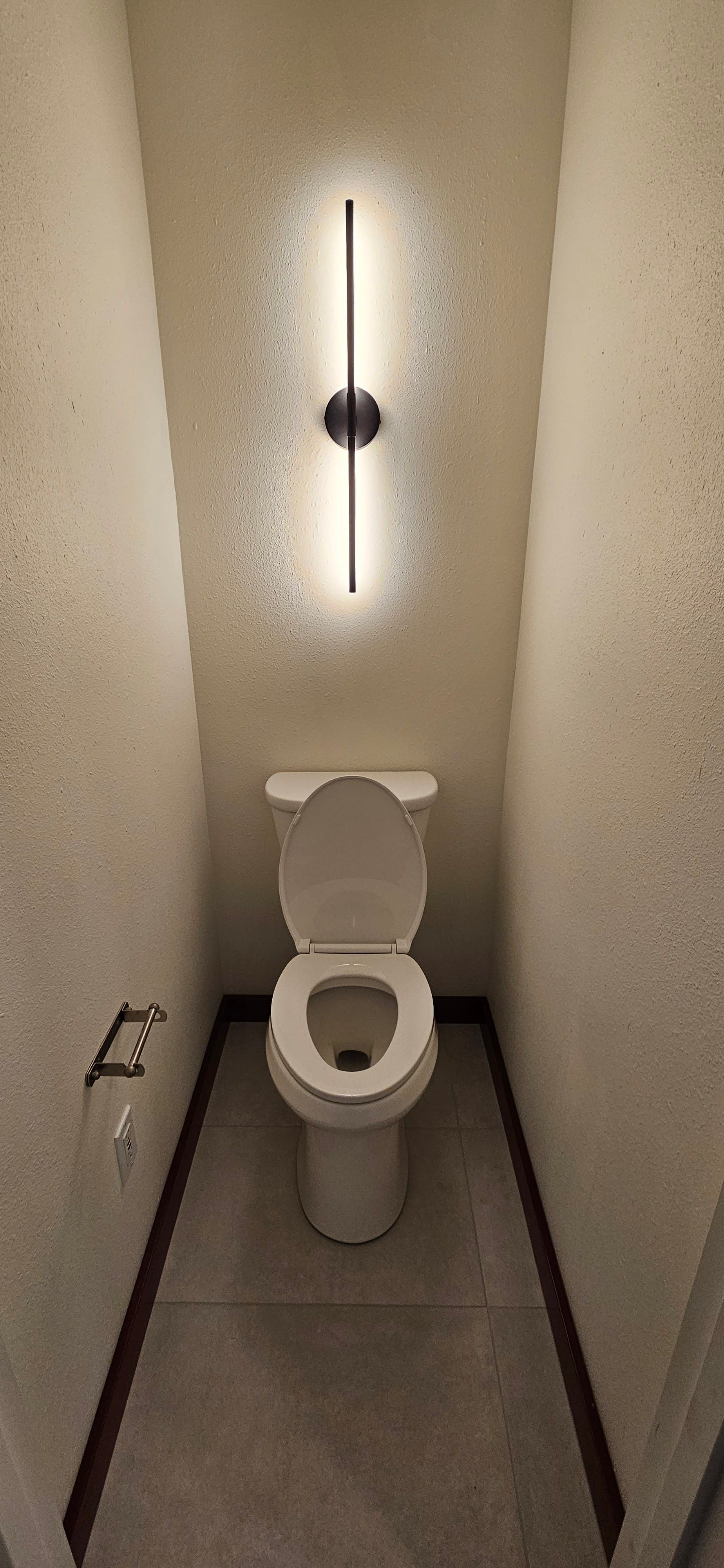A small bathroom with a toilet, and a vertical light fixture centered above it. The walls are speckled.