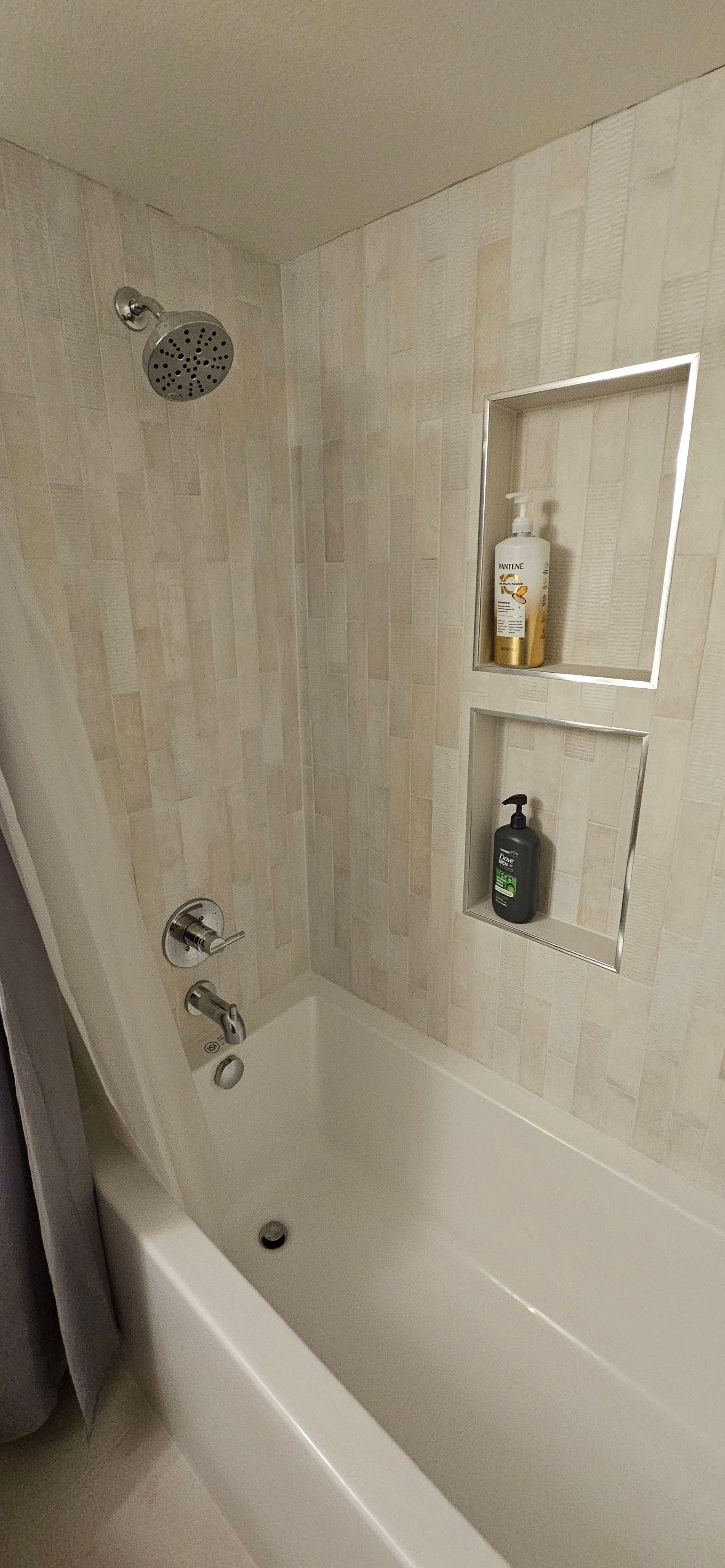 A small, white bathroom with light beige tiled walls, a showerhead, and a built-in shelf holding bottles.