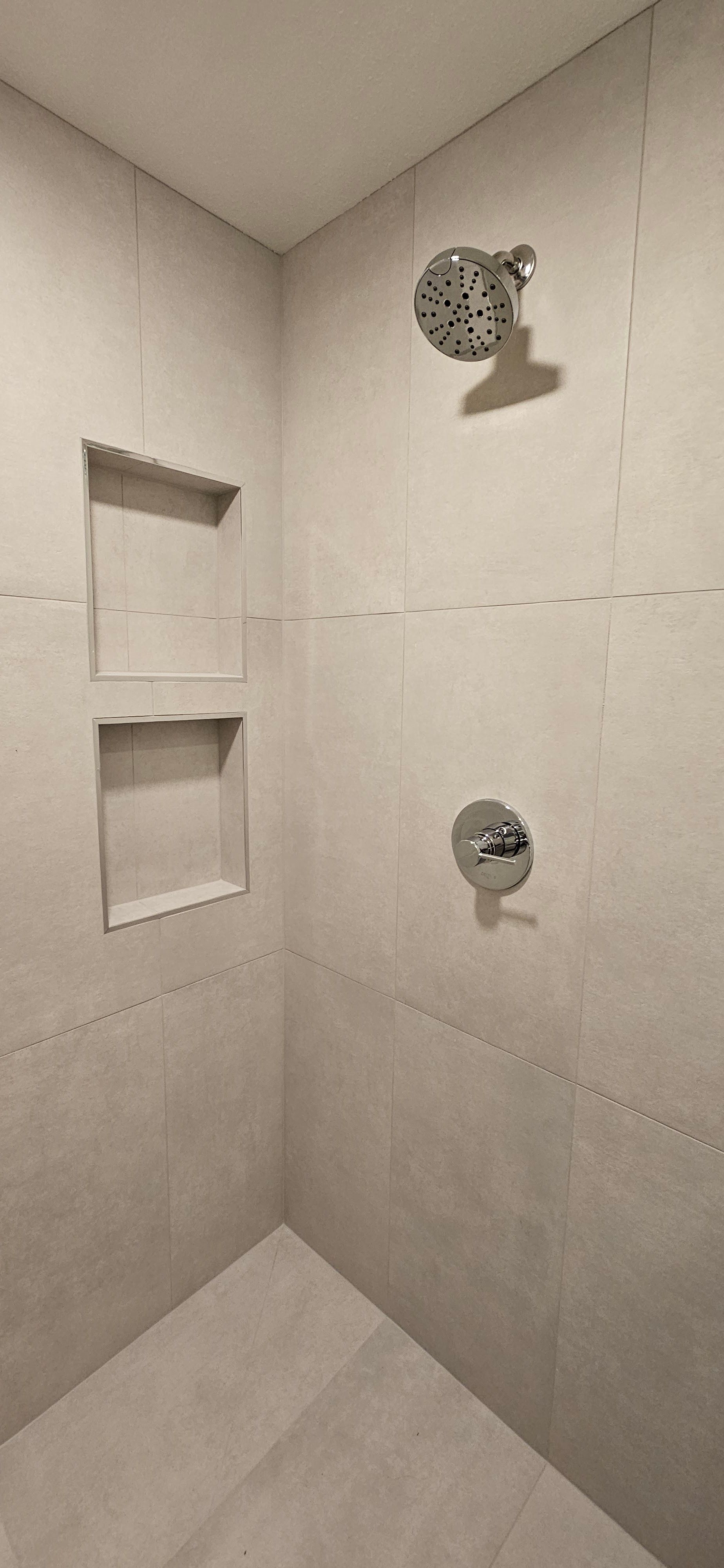 Corner shower with neutral-toned tiled walls and floor. Chrome shower head and controls are visible.