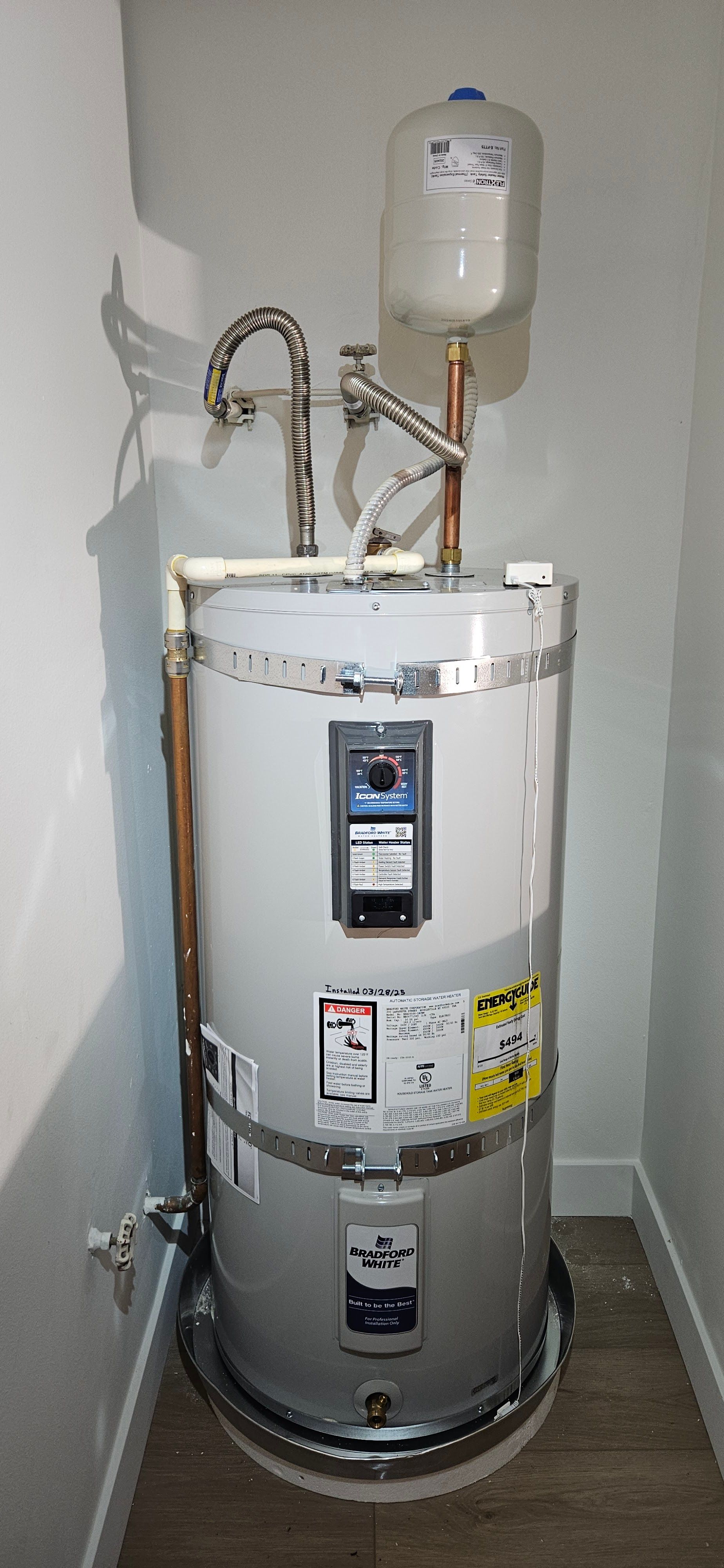 A water heater in a narrow closet. It is cylindrical and white with various pipes attached.