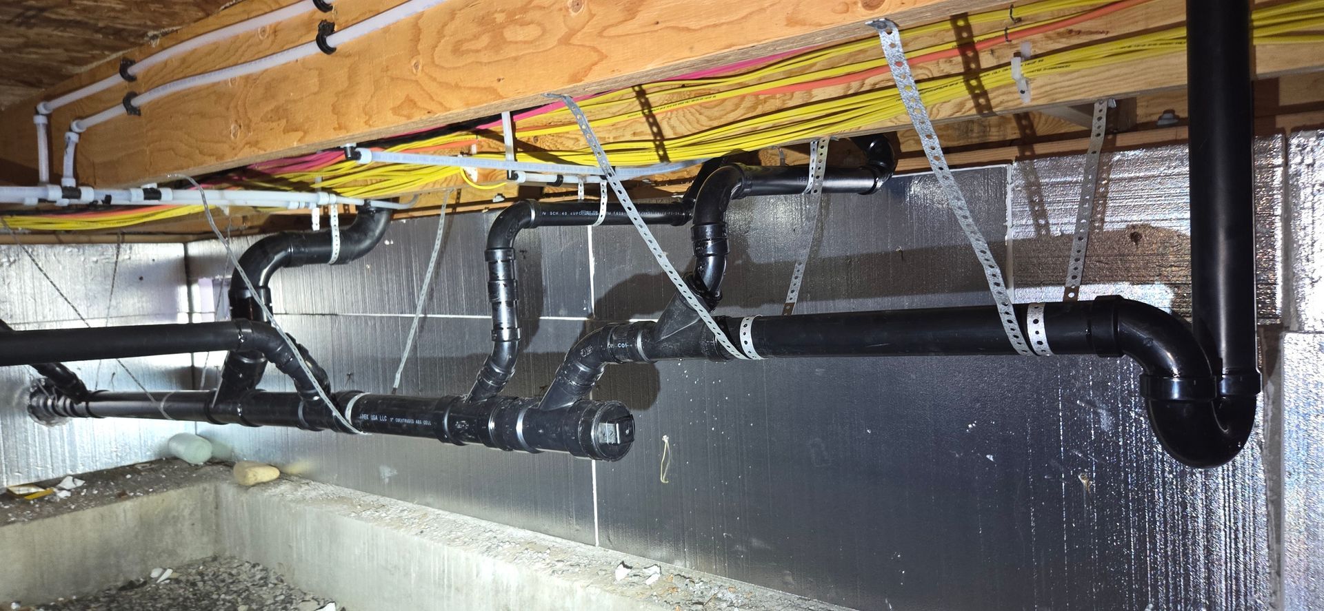 Black plumbing is suspended from the ceiling. Insulation is on the wall behind the pipes.