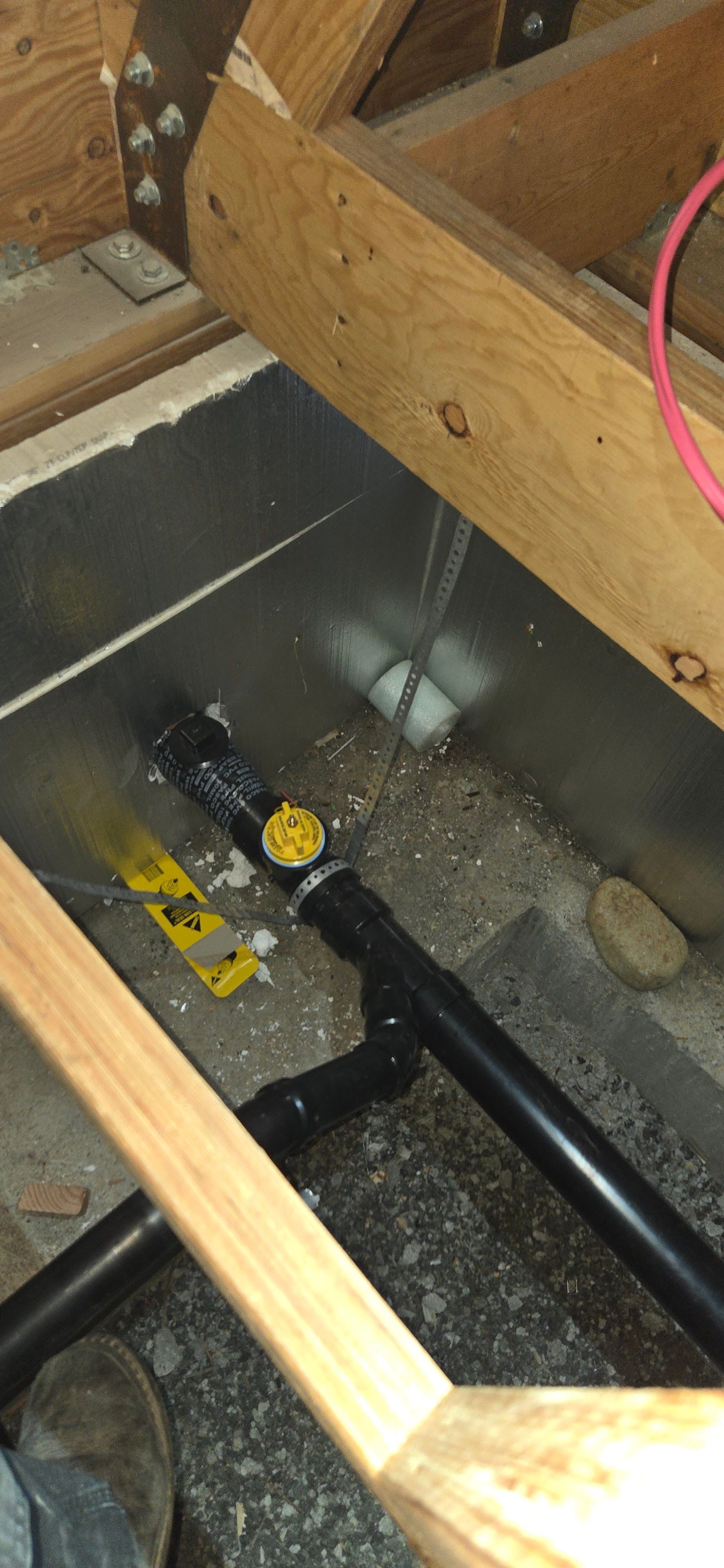 Plumbing pipes under wooden beams. A black pipe with a yellow valve and other joints. Grey and white flooring.