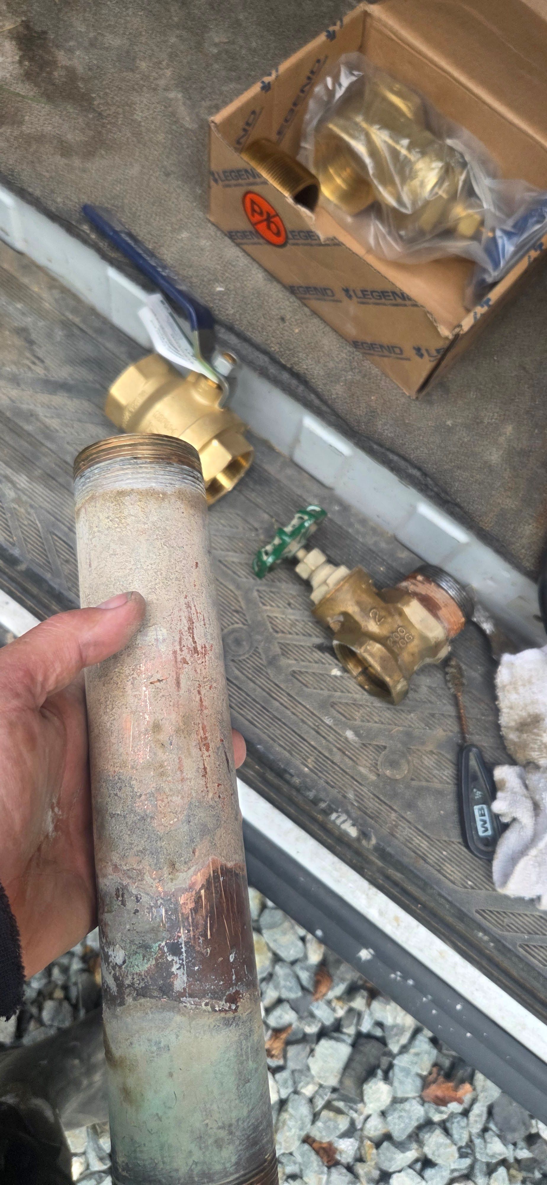 A hand holds a corroded pipe next to new valves, a box, and tools, likely for plumbing work.