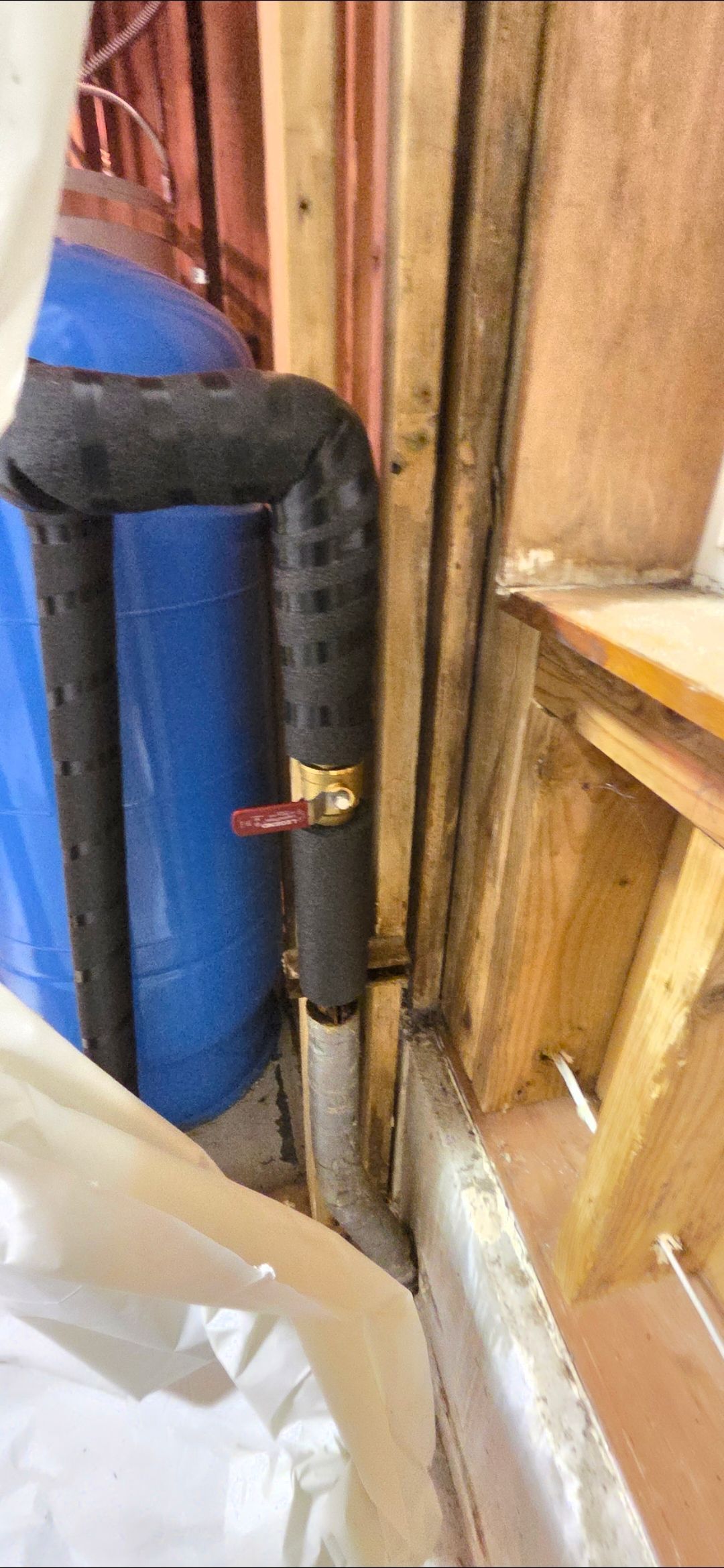 Black-insulated pipes with a red valve near a blue tank and wooden framing.