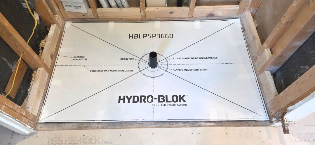 A white HYDRO-BLOK shower base in a wood-framed bathroom. Black drain pipe in the center.