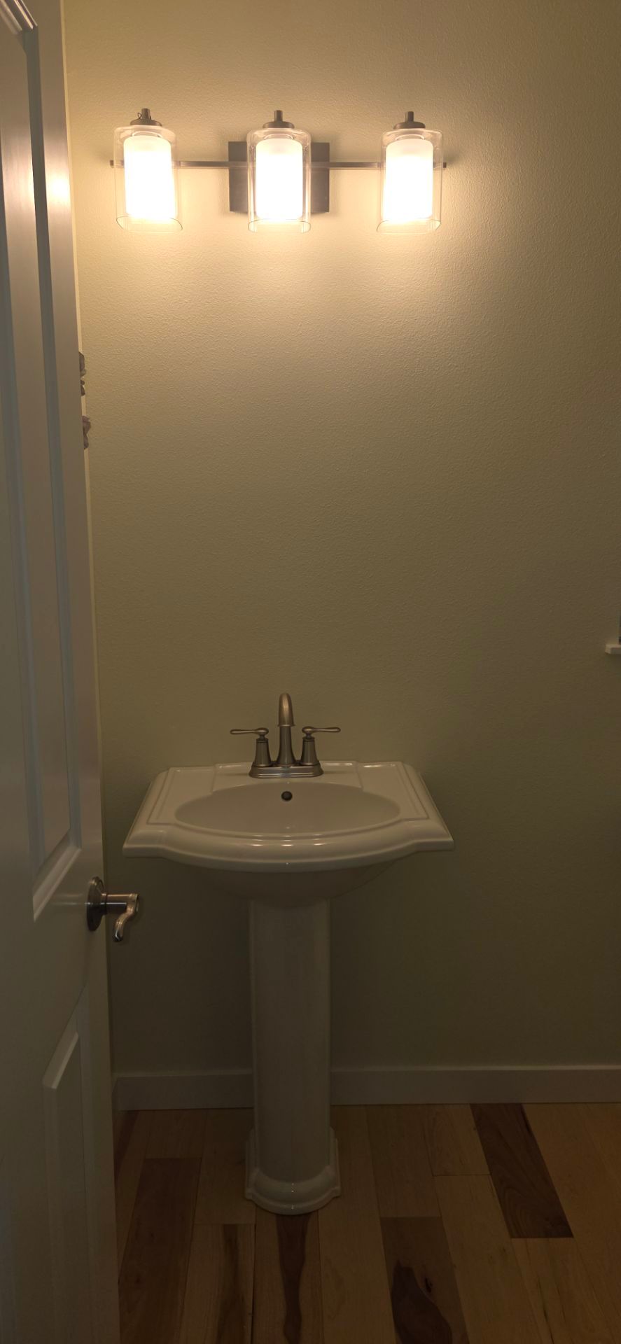 Bathroom with pedestal sink, three-light vanity, door on left, and hardwood floor.