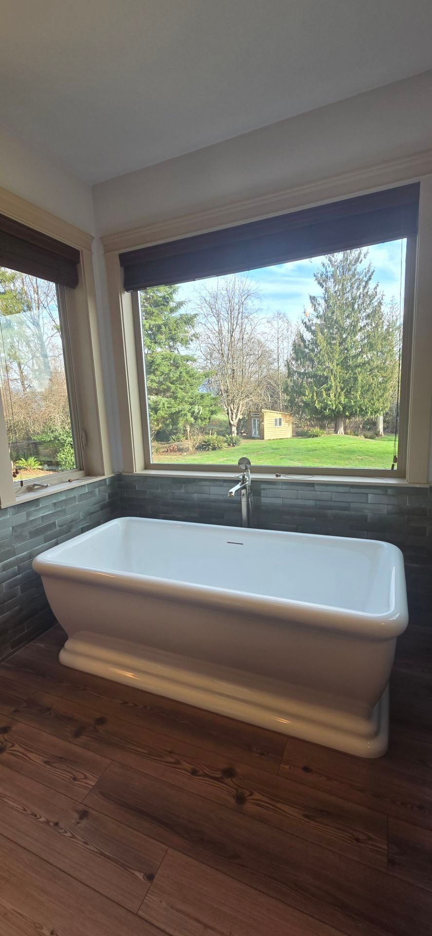 White bathtub by a window overlooking a green yard and trees. Wooden floor.