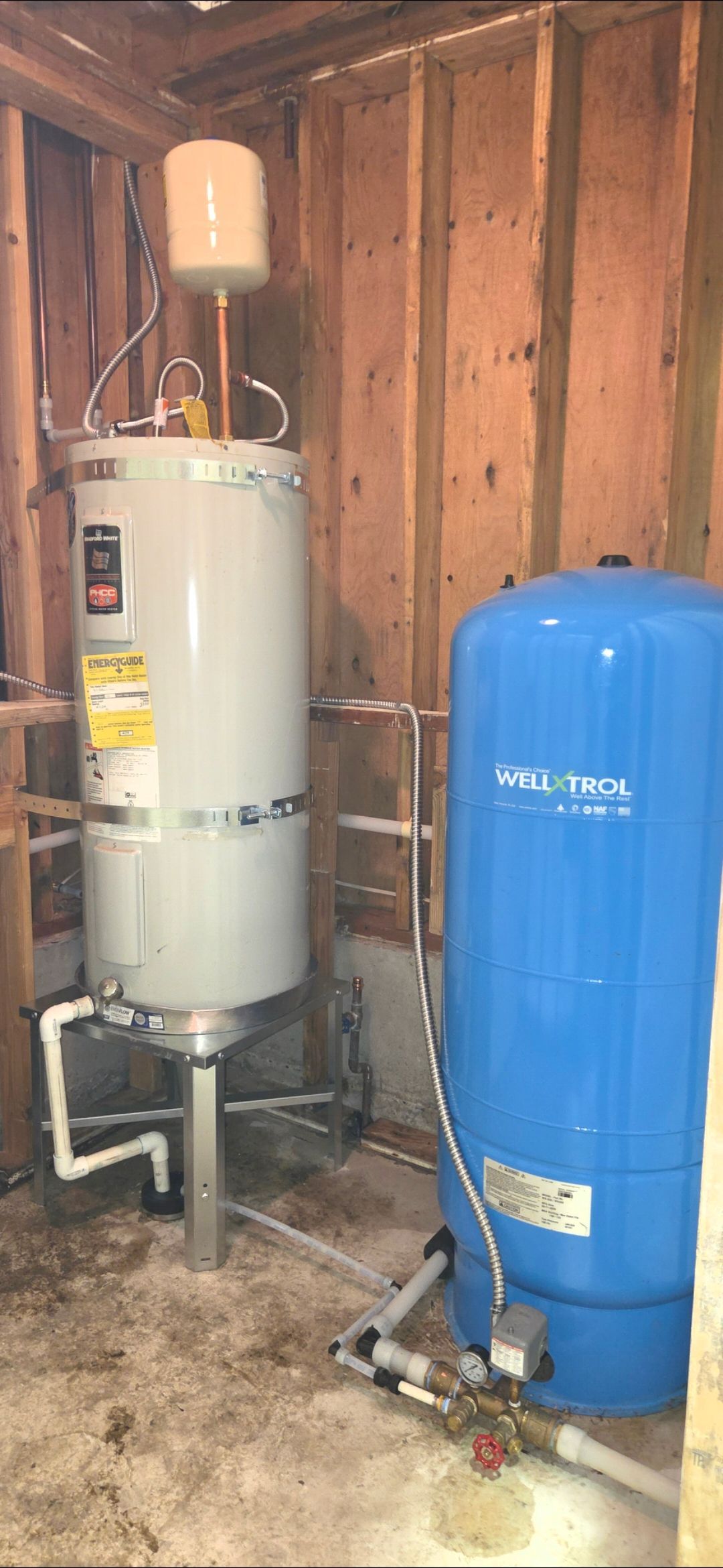 A water heater and a blue water tank in a room with wood walls and a concrete floor.