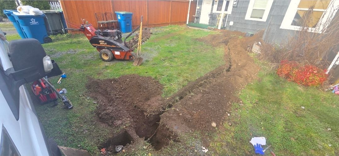 A yard with a trench being dug, a snowblower, and trash cans near a house.