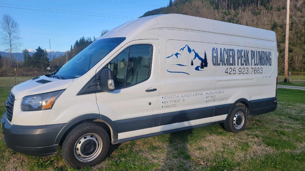 White plumbing service van with logo parked on grass in front of trees and mountains.