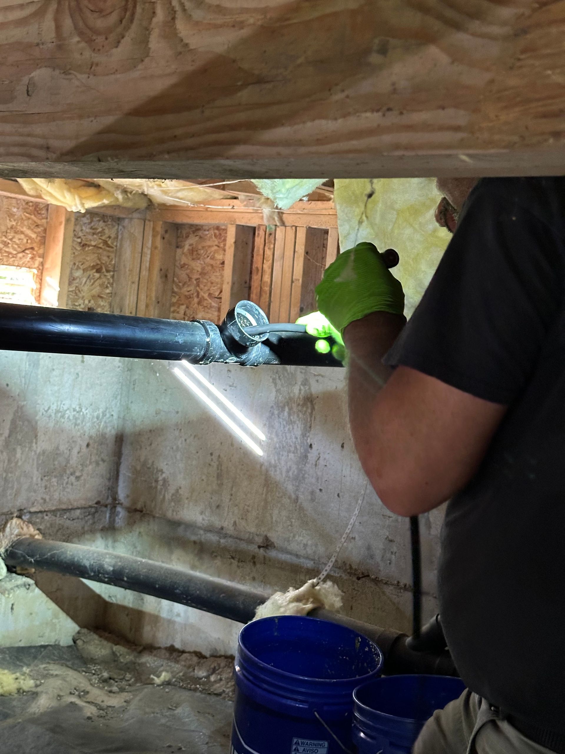 Person inspecting pipes in a crawl space, using a flashlight and wearing green gloves.