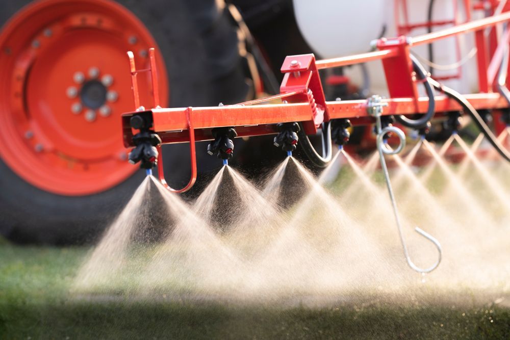 Agricultural sprayer applying liquid chemicals to a field; orange tractor wheel visible.