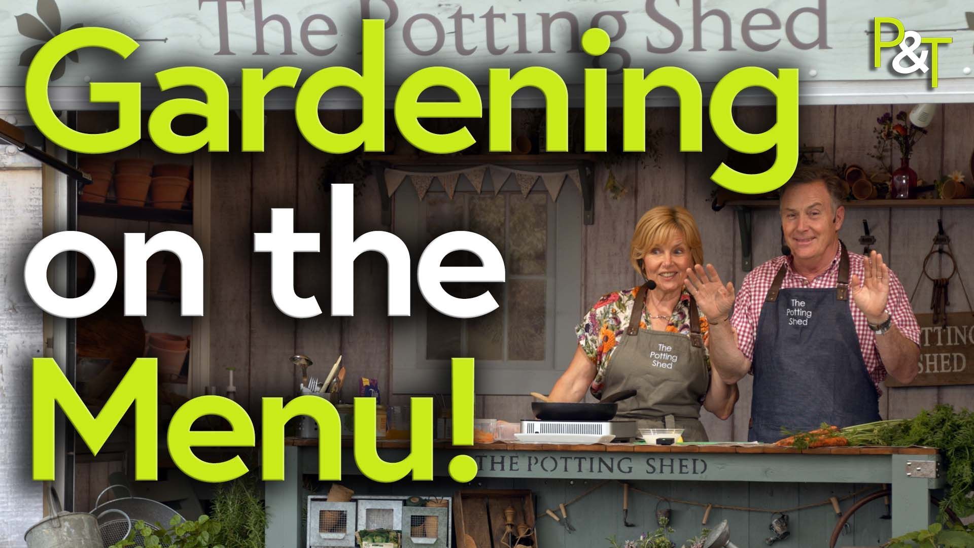 Martin and Jill Fish behind a counter on a stage with the sign above them reading The Potting Shed Stage. They are demonstrating some cookery and there's large green and white text over them saying Gardening on the Menu.
