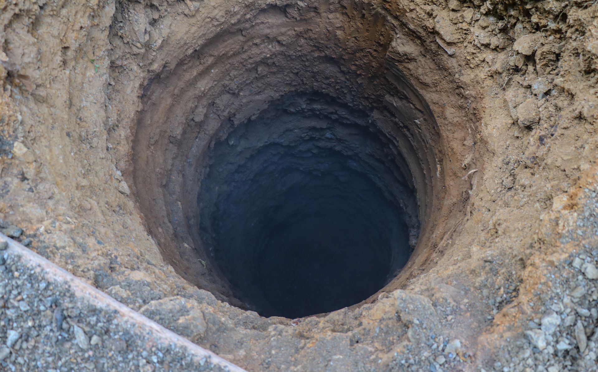 Deep, dark hole in the ground, viewed from above; dirt and rock surround the opening.