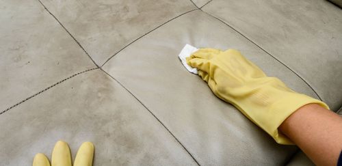Person cleaning a leather couch with a yellow-gloved hand, using a white wipe.