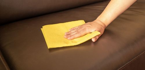 A person's hand wiping a yellow cloth on a brown leather couch.