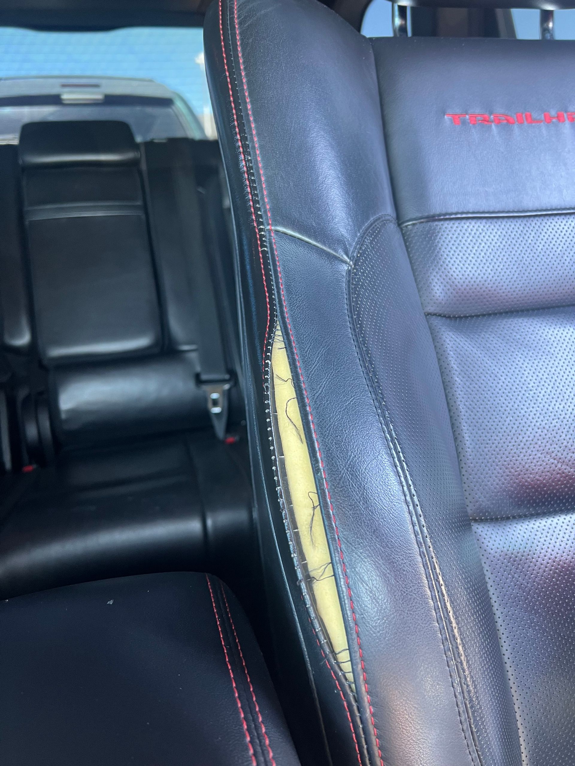 Close-up of a damaged black leather car seat with a large tear exposing yellow foam. Red stitching is visible.