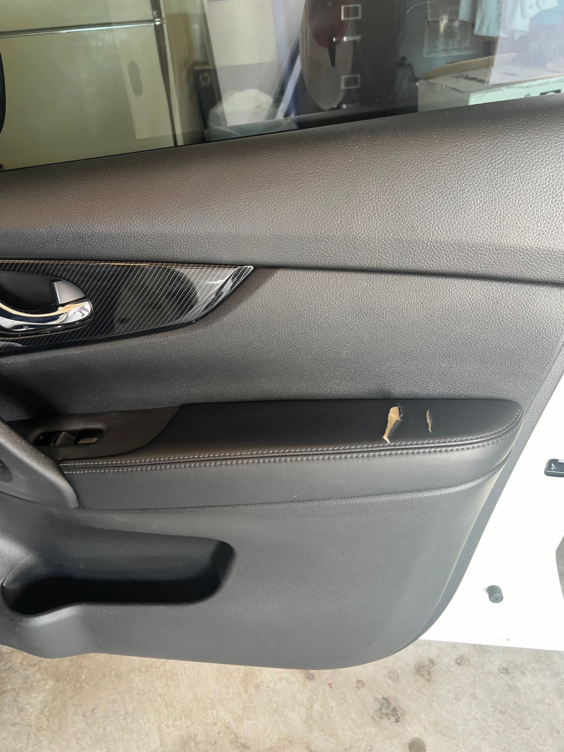 Black car door panel, worn leather armrest, carbon fiber detail, window visible.