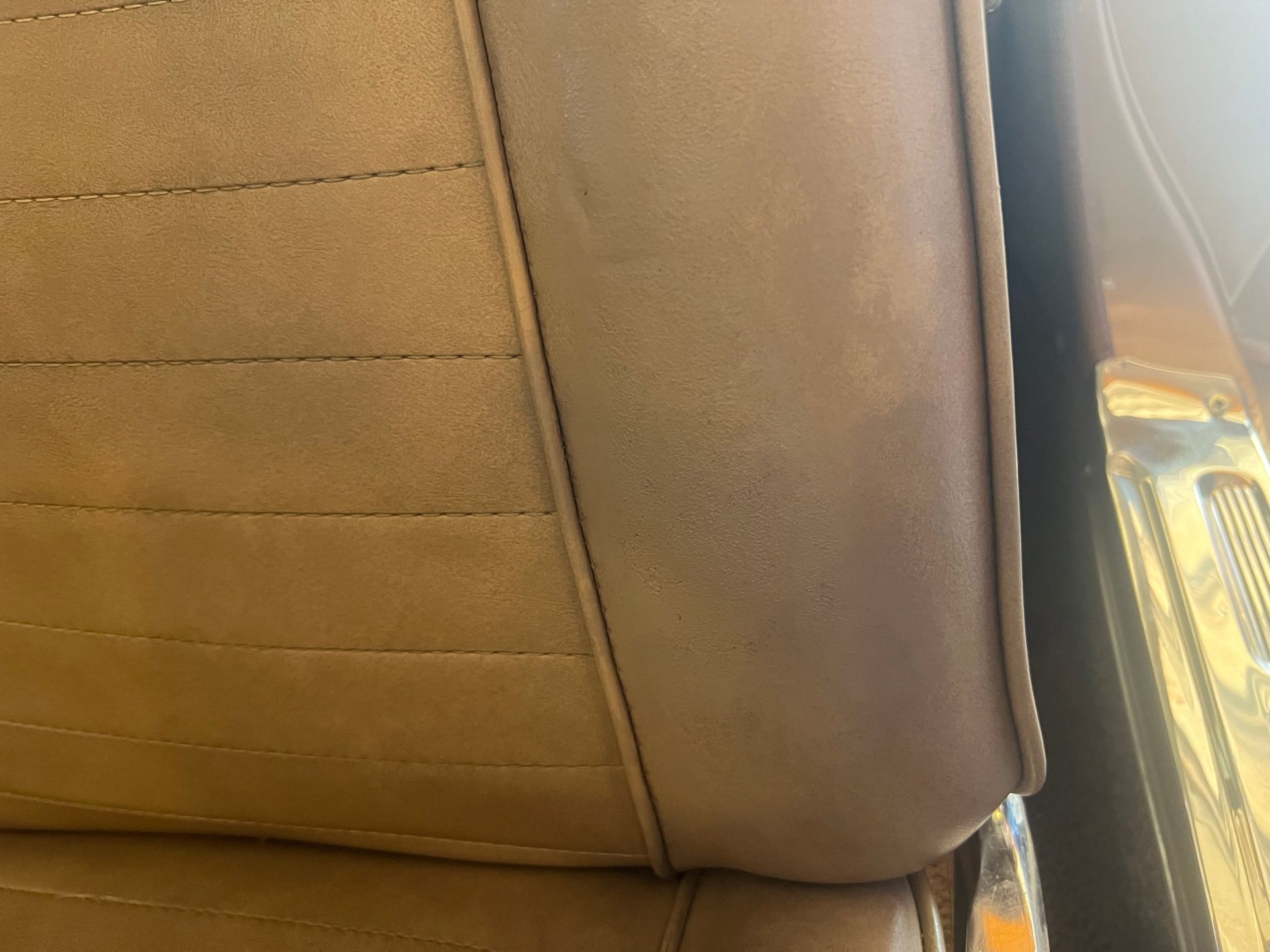 Tan car seat with stitched horizontal lines and a smooth, dark-tan border next to a metal panel.