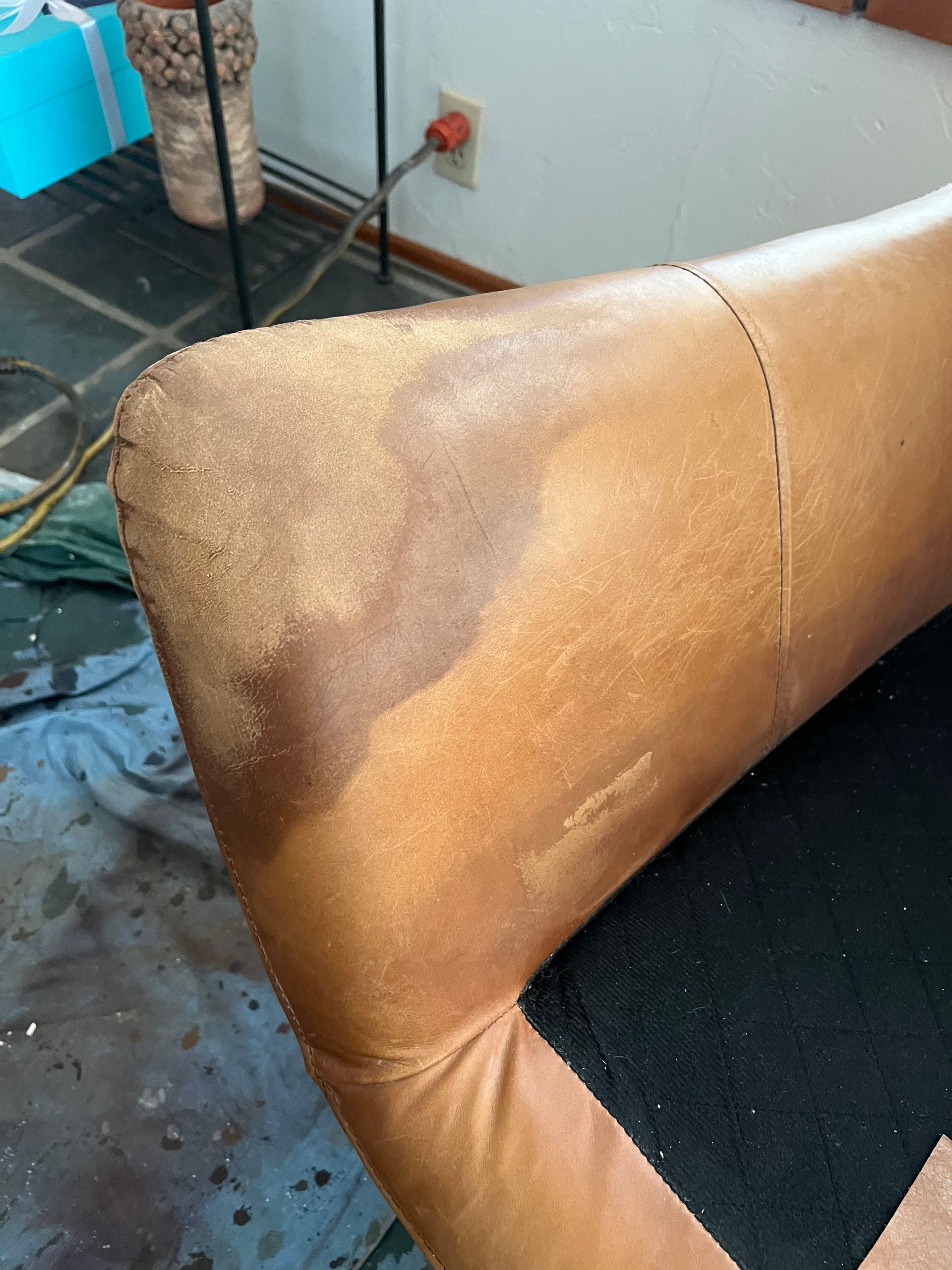 Close-up of a leather couch corner showing color variations and minor damage in a room with a brick floor.