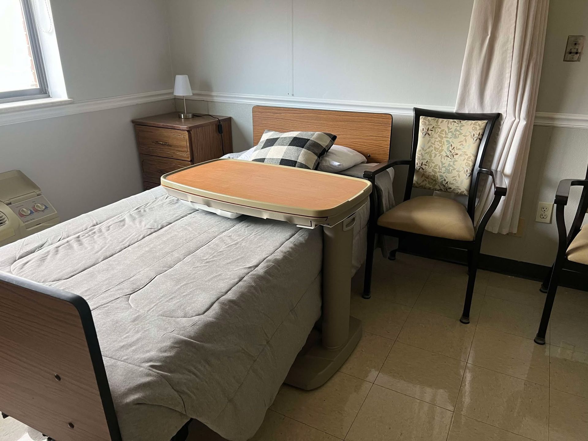 A hospital room with a bed , chair , table and window.