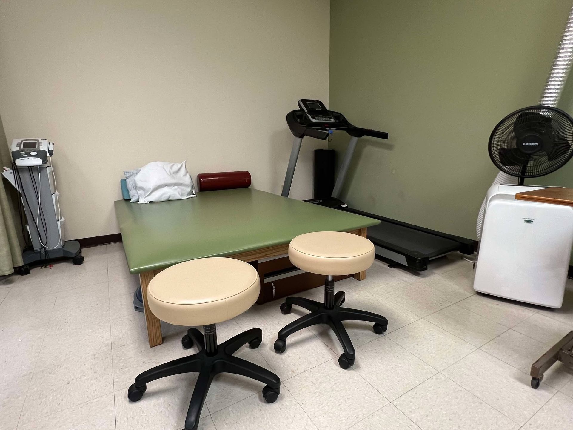 A room with a table , stools , a treadmill and a fan.