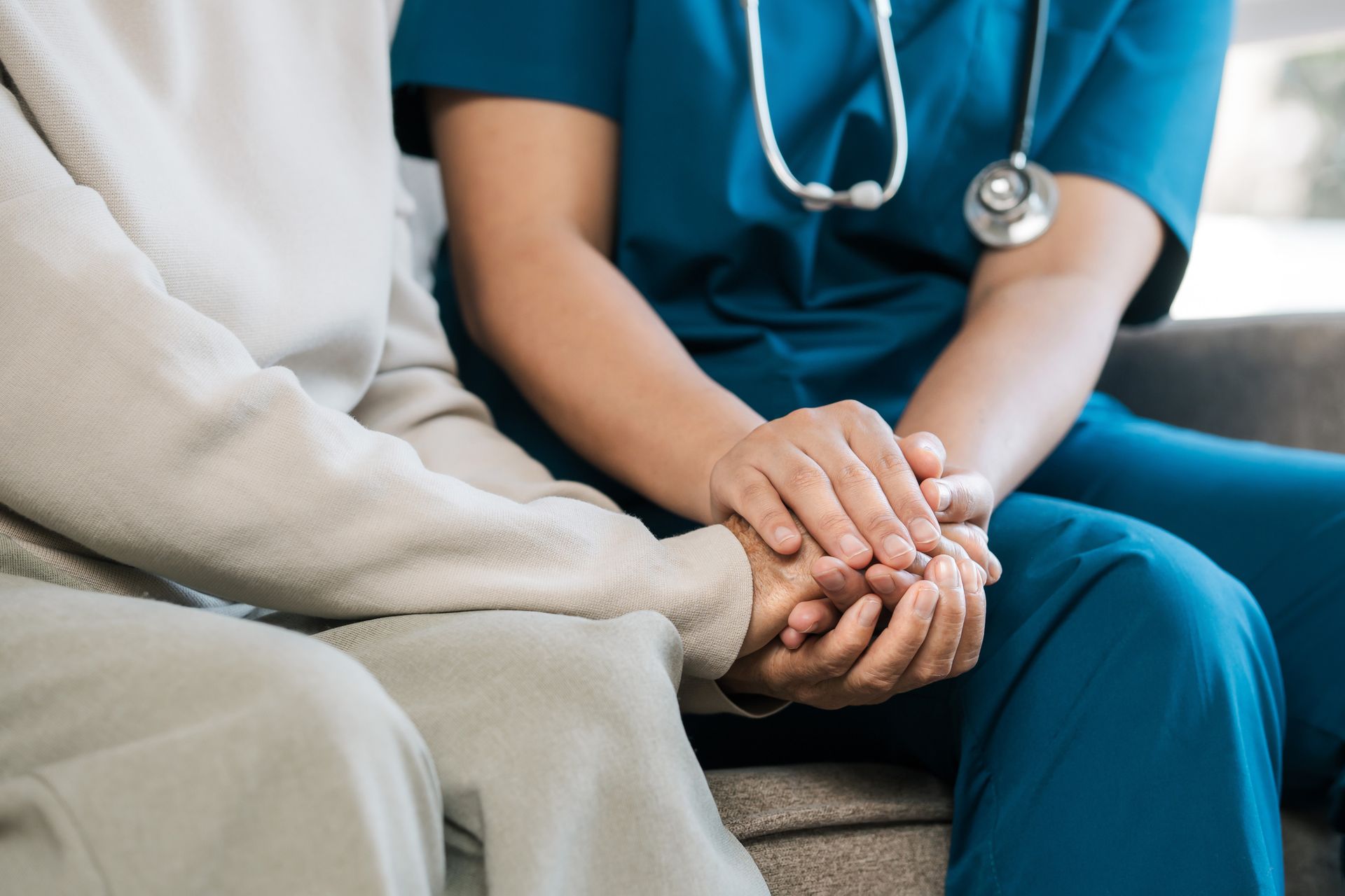 A nurse is holding the hand of an elderly woman while sitting on a couch.
