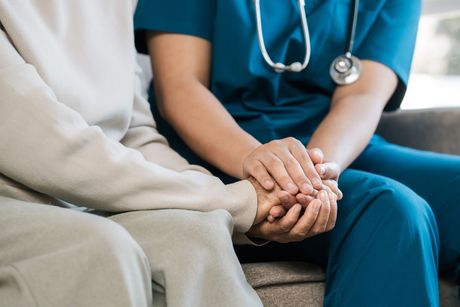 A nurse is holding the hand of an elderly woman while sitting on a couch.