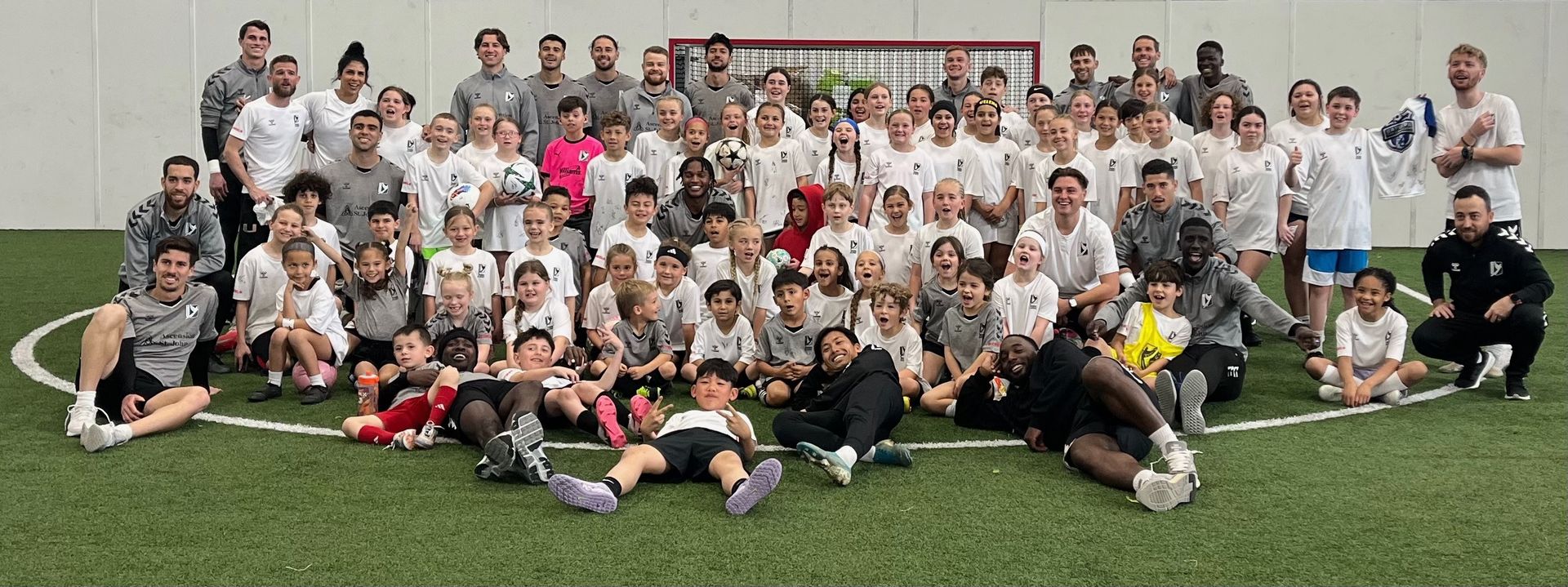 Group of people posing on a green soccer field, mostly wearing white shirts.
