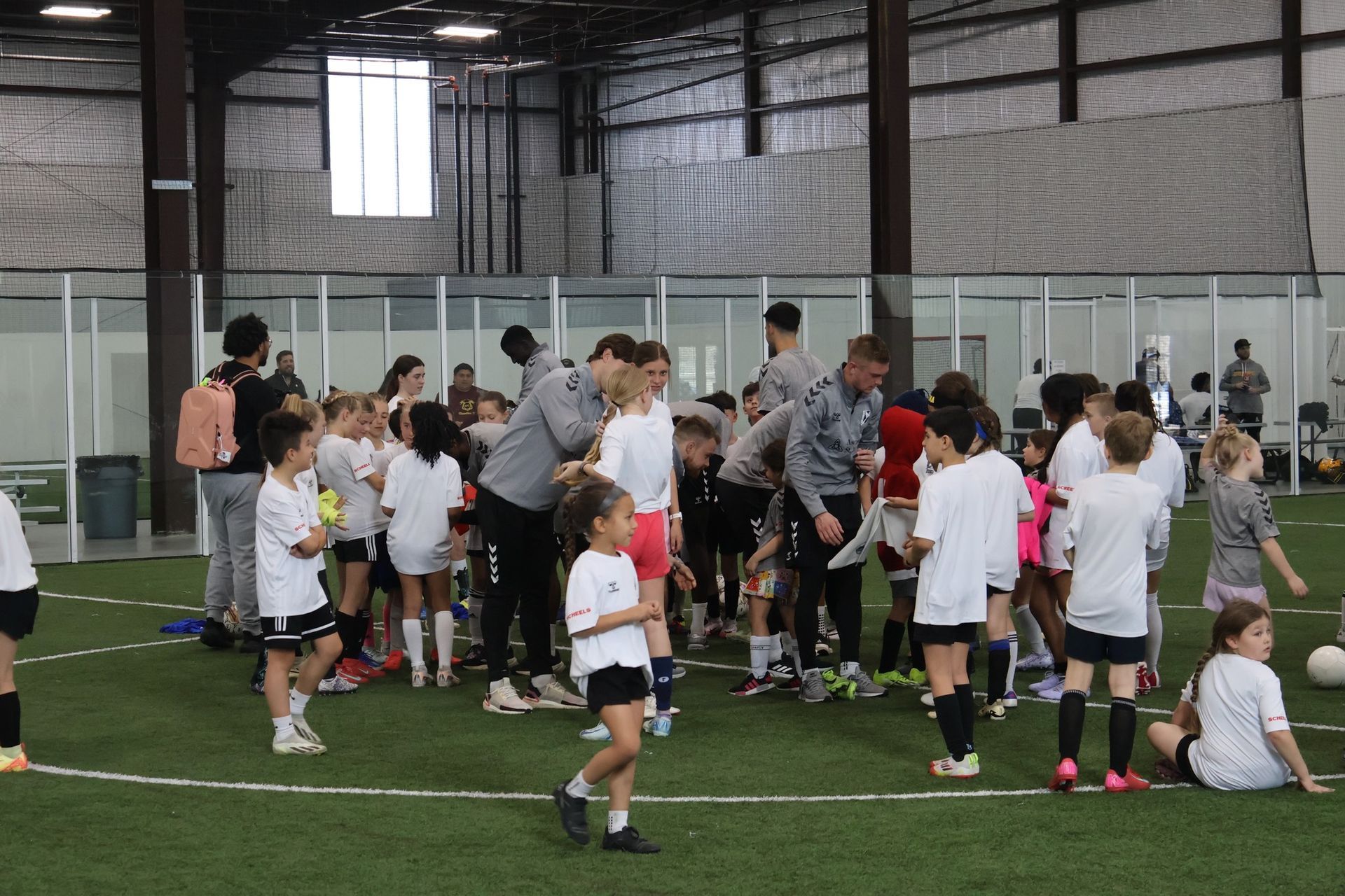 Children in white soccer jerseys and shorts on an indoor field with coaches.