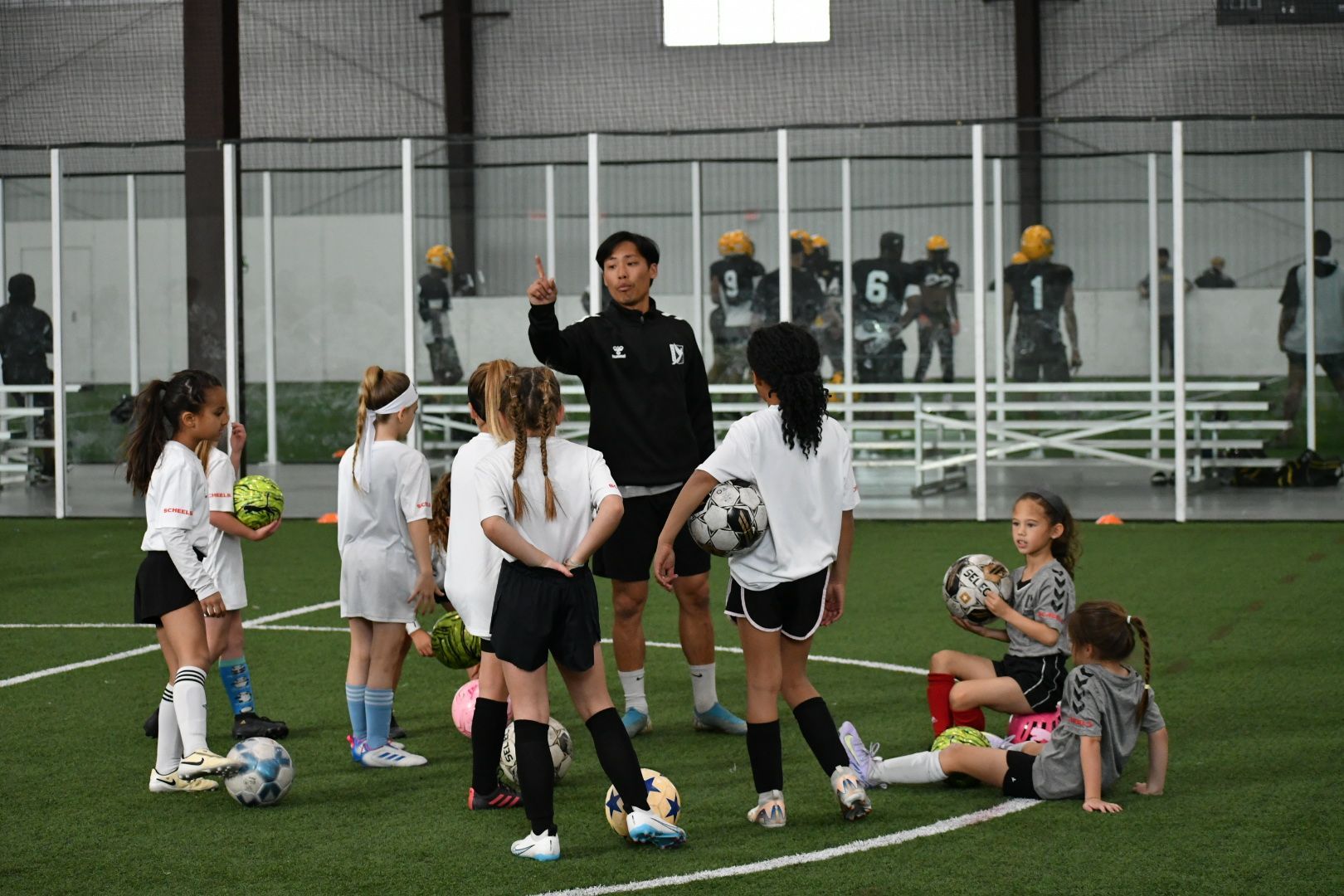 A soccer coach instructs a group of young girls on an indoor field.