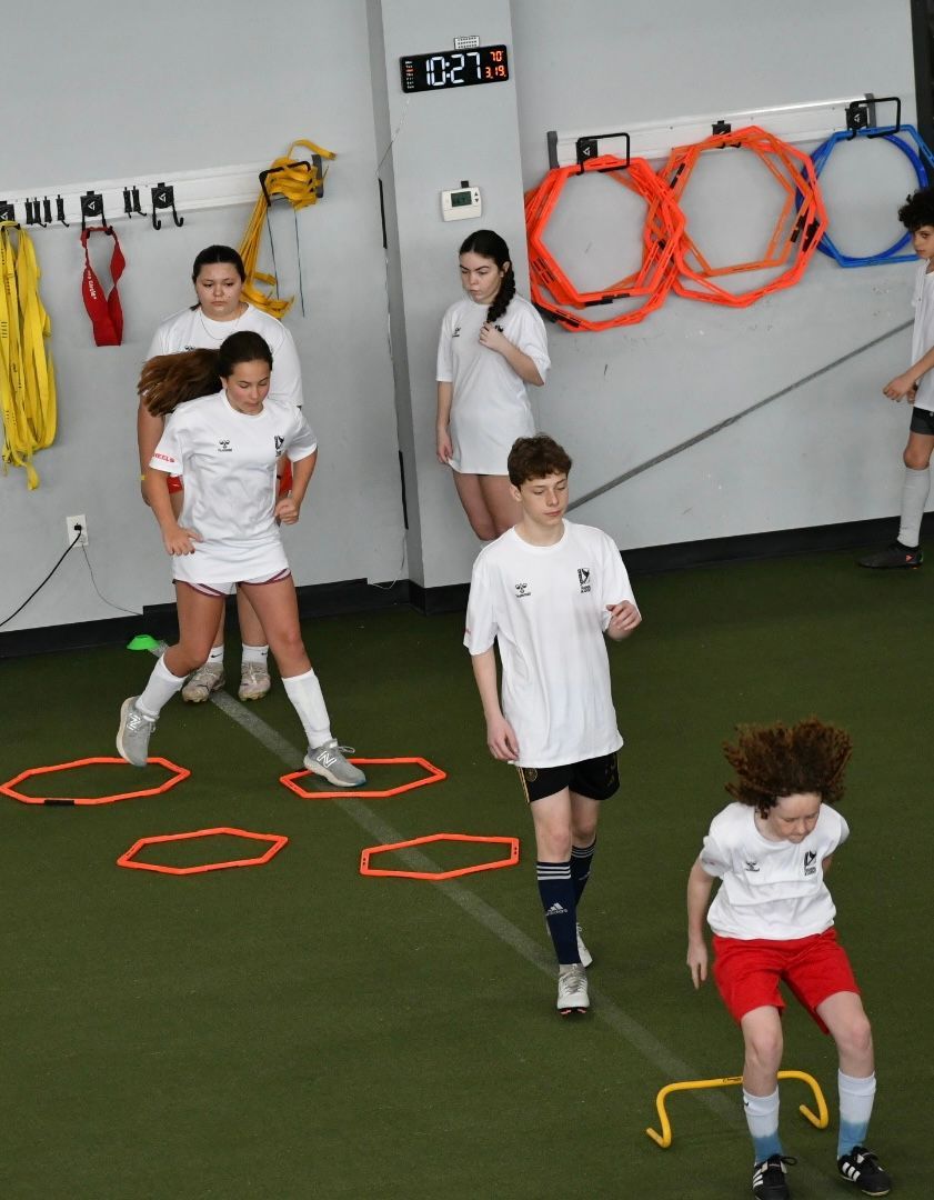 Group of young athletes in a gym, practicing agility drills with cones and hurdles.