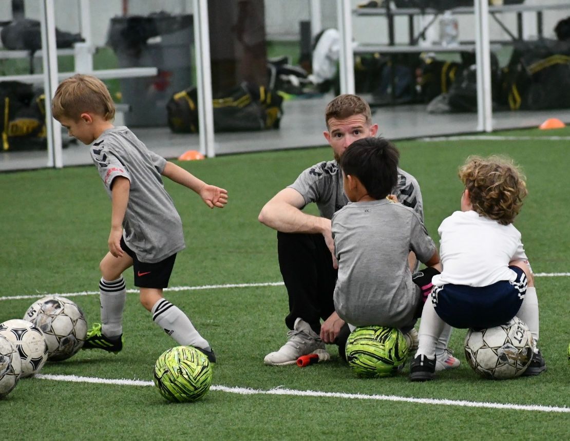 Coach teaching young children soccer on a green field indoors.