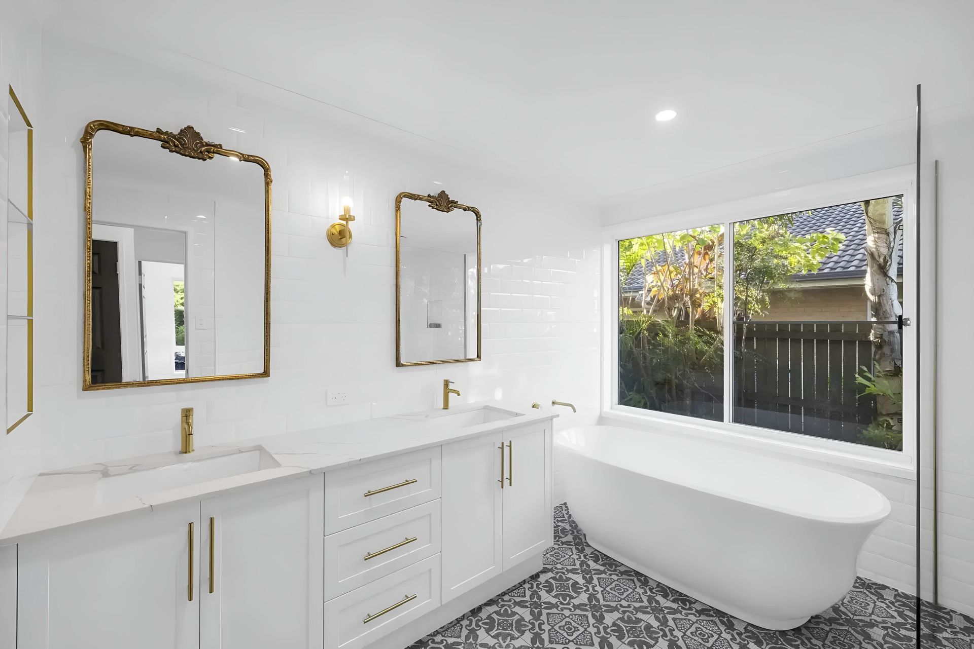 White bathroom with gold accents: double vanity, mirrors, freestanding tub, window with a view, patterned floor — CK Cabinetry in Coolum Beach, QLD